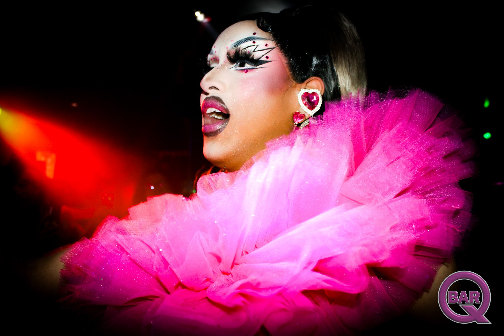 Close-up of a drag performer with elaborate makeup and a pink feather costume, performing on stage in a dark setting with colorful lights.