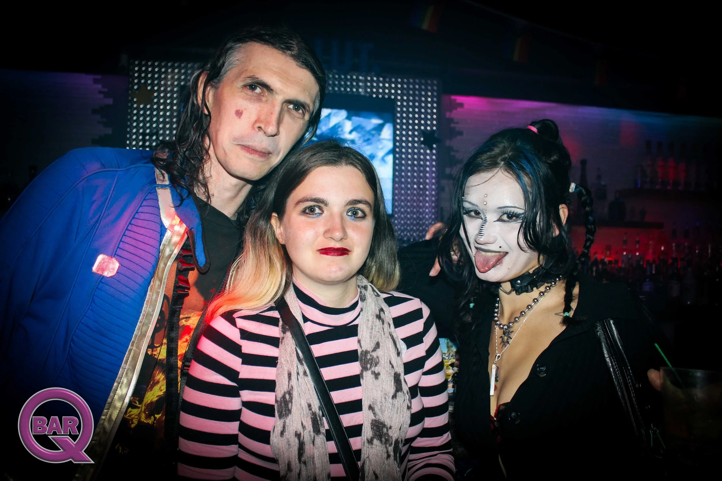 Three people at a nightclub, two women and one man, posing together with colorful lighting and a bar sign visible in the background.