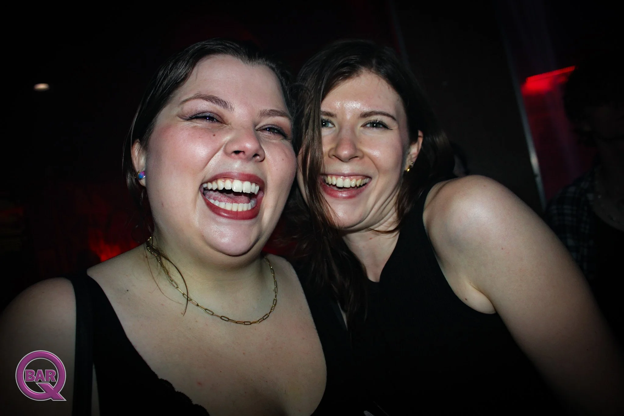Two smiling women taking a selfie in a dark setting, possibly a bar or nightclub, with one woman in the foreground and the other behind her.