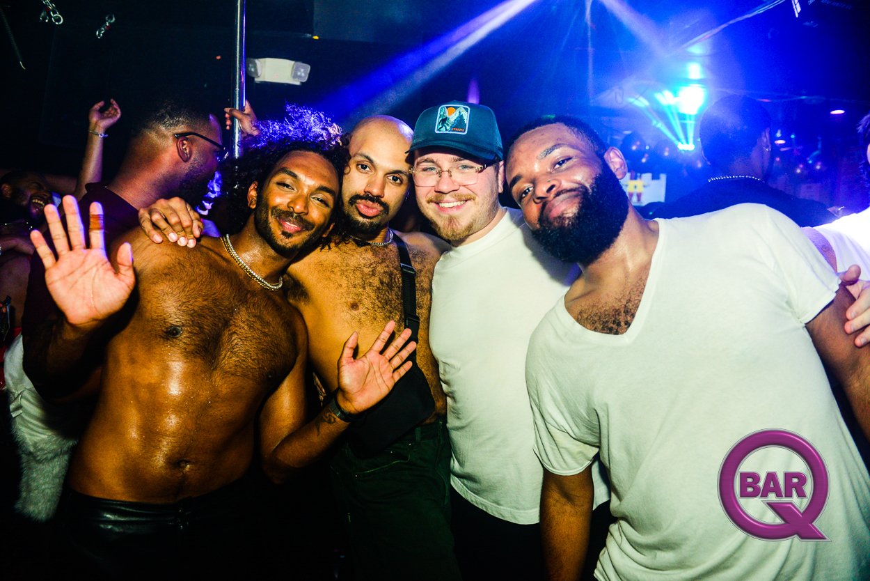 Four men smiling and posing together in a nightclub with colorful lights. Two men are shirtless, and the others are wearing white t-shirts. One of the shirtless men is waving, and the group appears to be having fun.