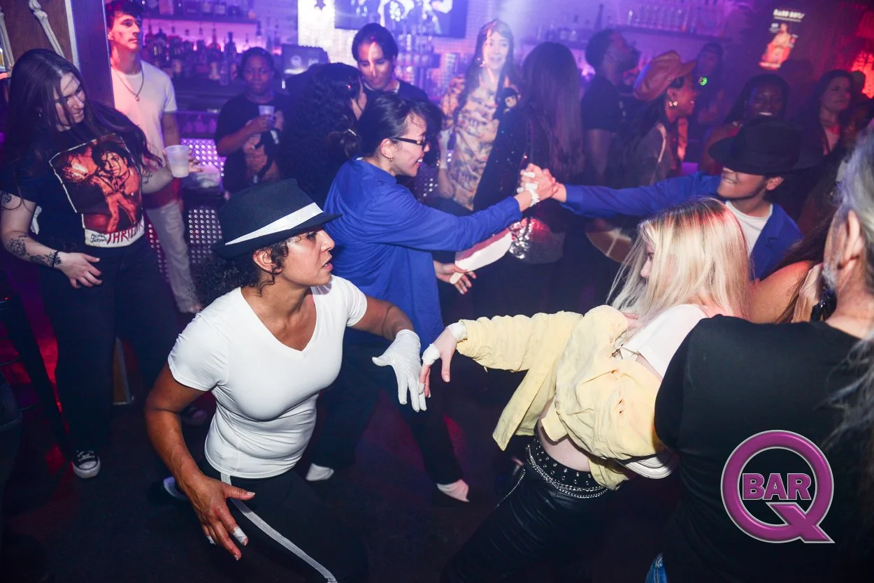People dancing and socializing at a lively bar or nightclub with colorful lighting and a city skyline backdrop. Some individuals are holding drinks, others are dancing in pairs or groups, and everyone appears to be enjoying the energetic atmosphere.