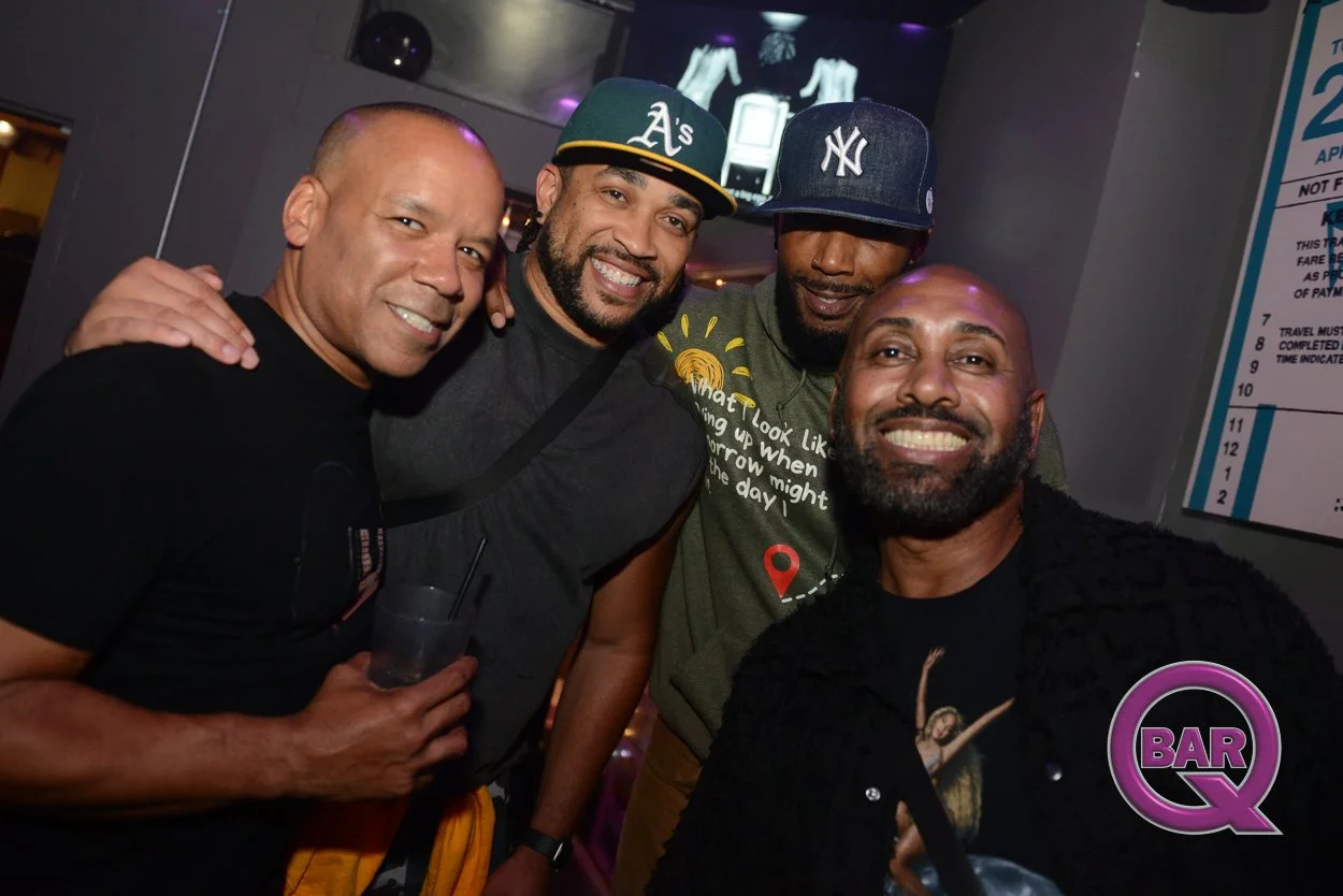Four men smiling and posing together in a bar or nightclub, with a TV screen and bar sign in the background.
