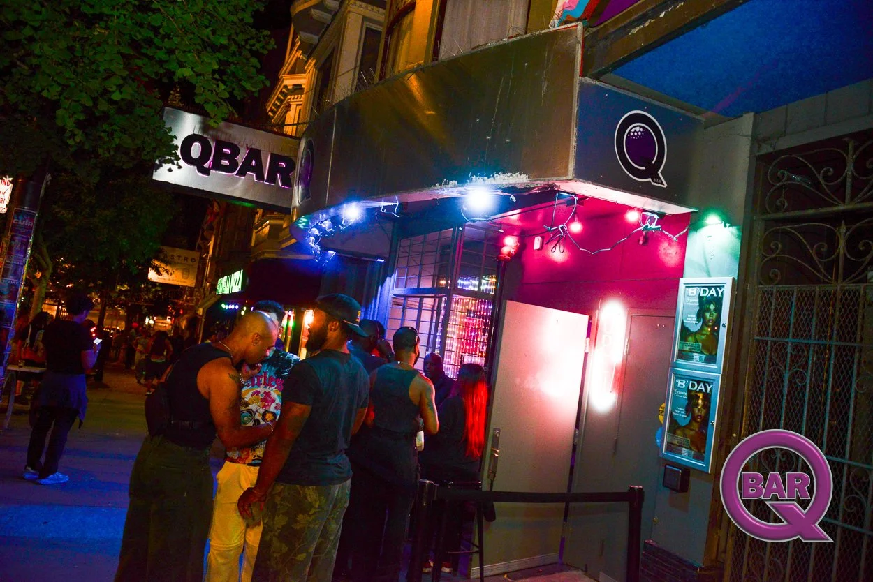 People standing outside a bar at night, illuminated by pink and blue neon lights, with a sign that reads 'Q BAR' and posters on the wall.