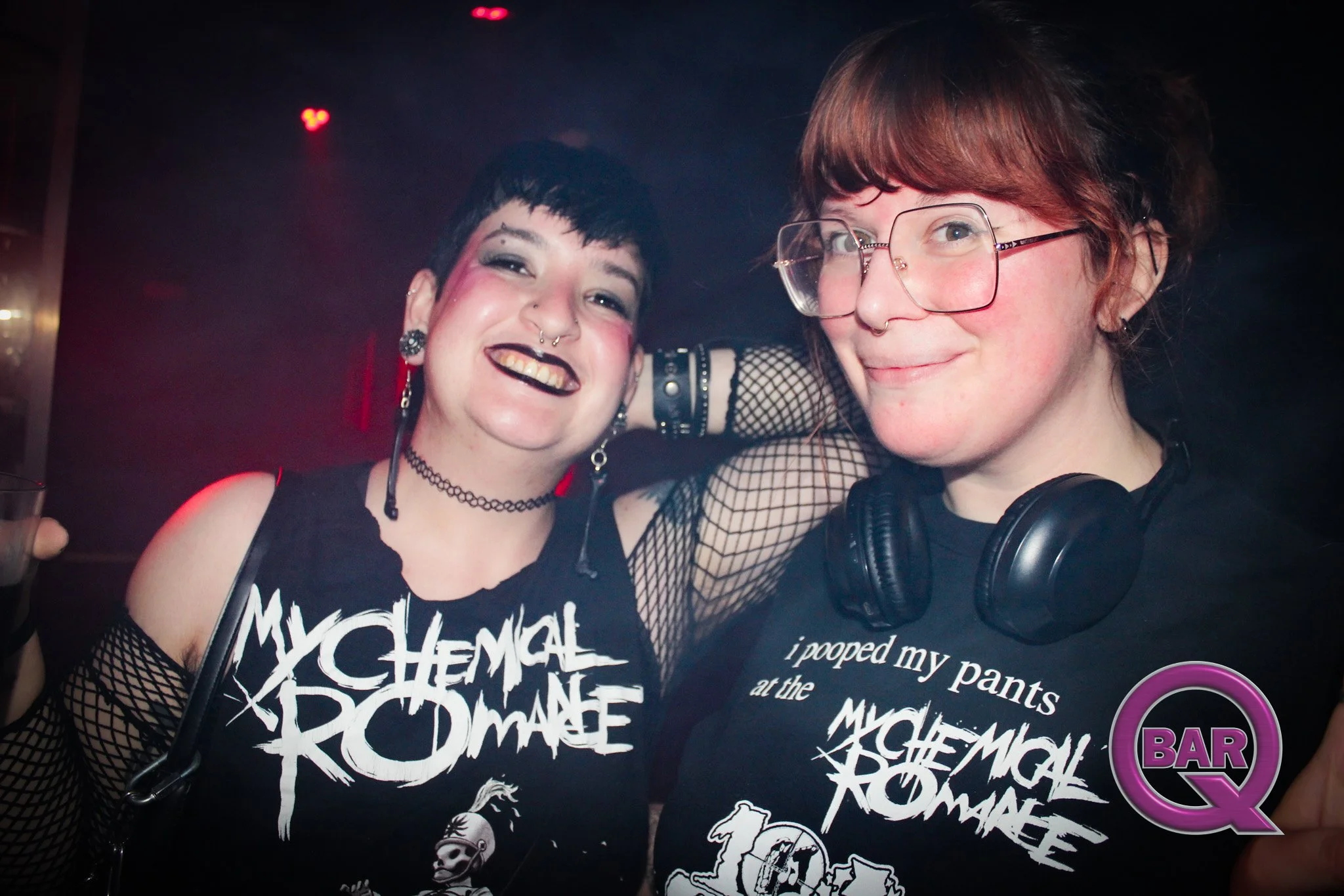 Two young women smiling in a nightclub, wearing black clothing and accessories, with one wearing glasses and the other with short dark hair, fishnet sleeves, and dark makeup.