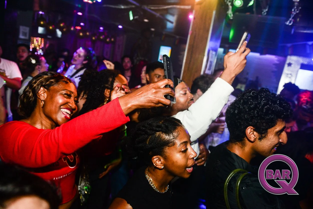 Crowd of people enjoying a night out at a bar or club, with some taking photos or videos, dancing, and smiling under colorful lighting.