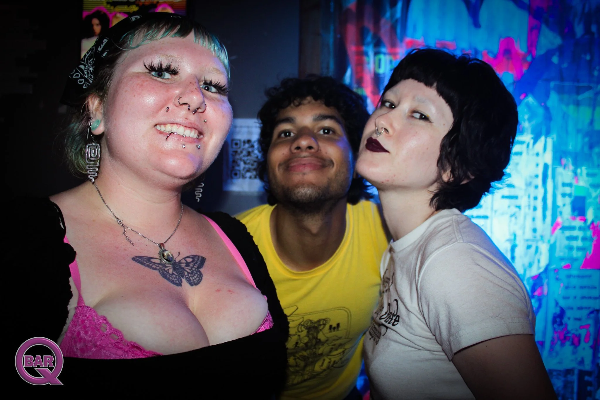 Three people taking a selfie at a nightclub with colorful neon lighting in the background, including a woman with green hair, a tattoo of a butterfly, and multiple facial piercings, alongside a man with curly black hair and a woman with short black h
