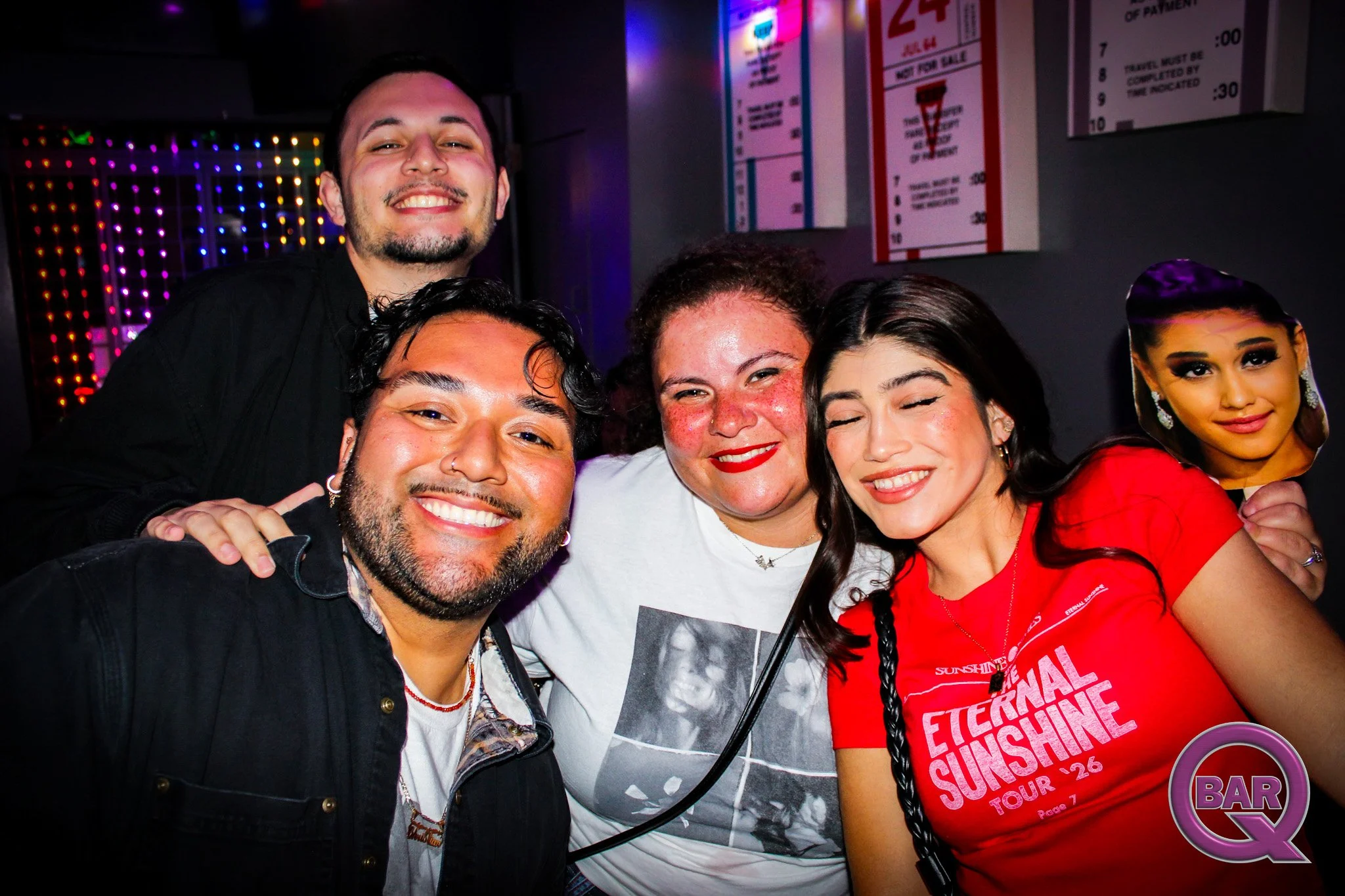 A group of five diverse friends, three women and two men, smiling and posing together at night in a bar with colorful neon signs and a menu on the wall behind them.