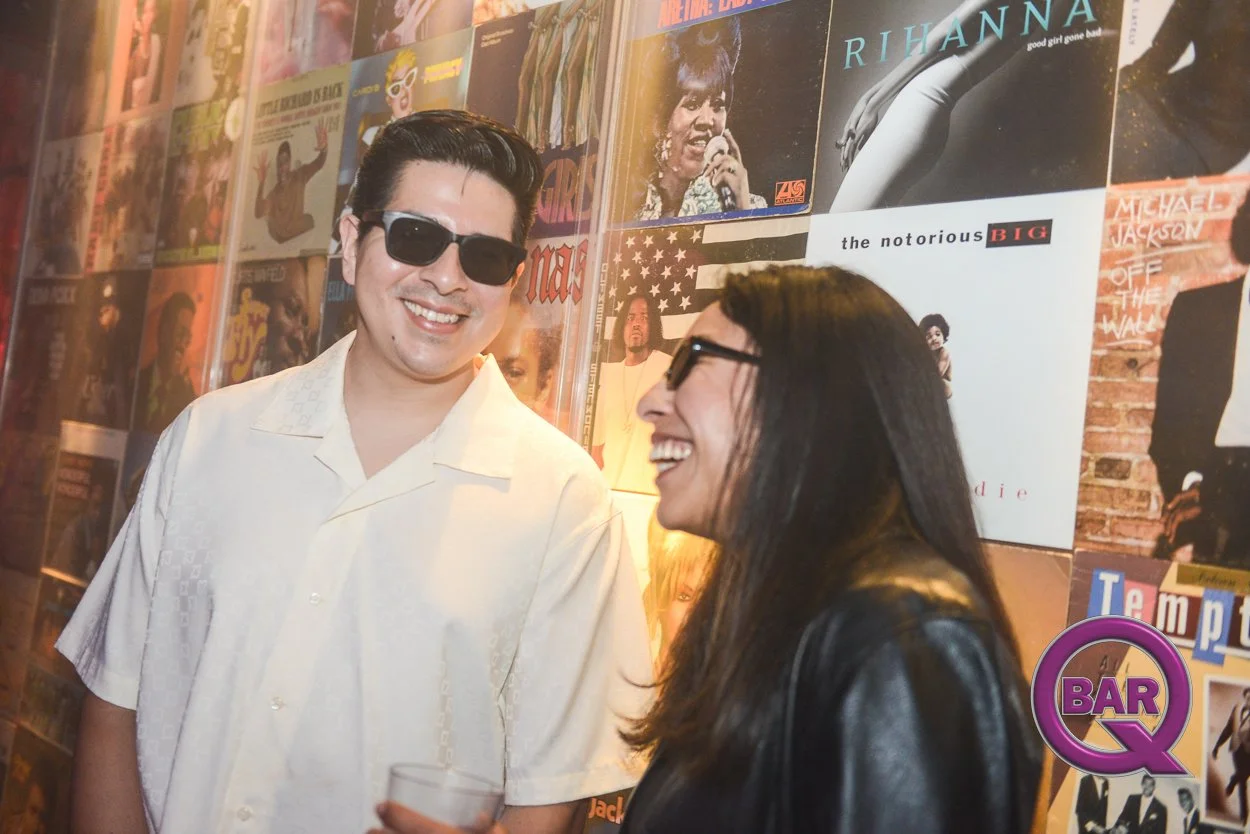 A man and woman smiling and laughing at a bar, with a wall of music album covers behind them. The man is wearing a white shirt and sunglasses. The woman has long dark hair and glasses, and is wearing a black jacket.