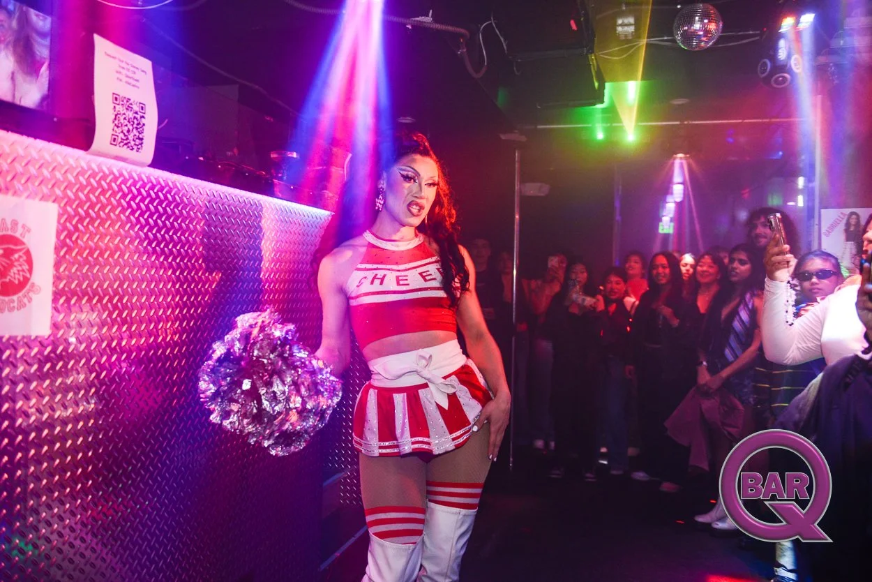 A drag performer dressed as a cheerleader with red and white outfit, holding silver pom-poms at a nightclub with colorful lights and an audience watching.
