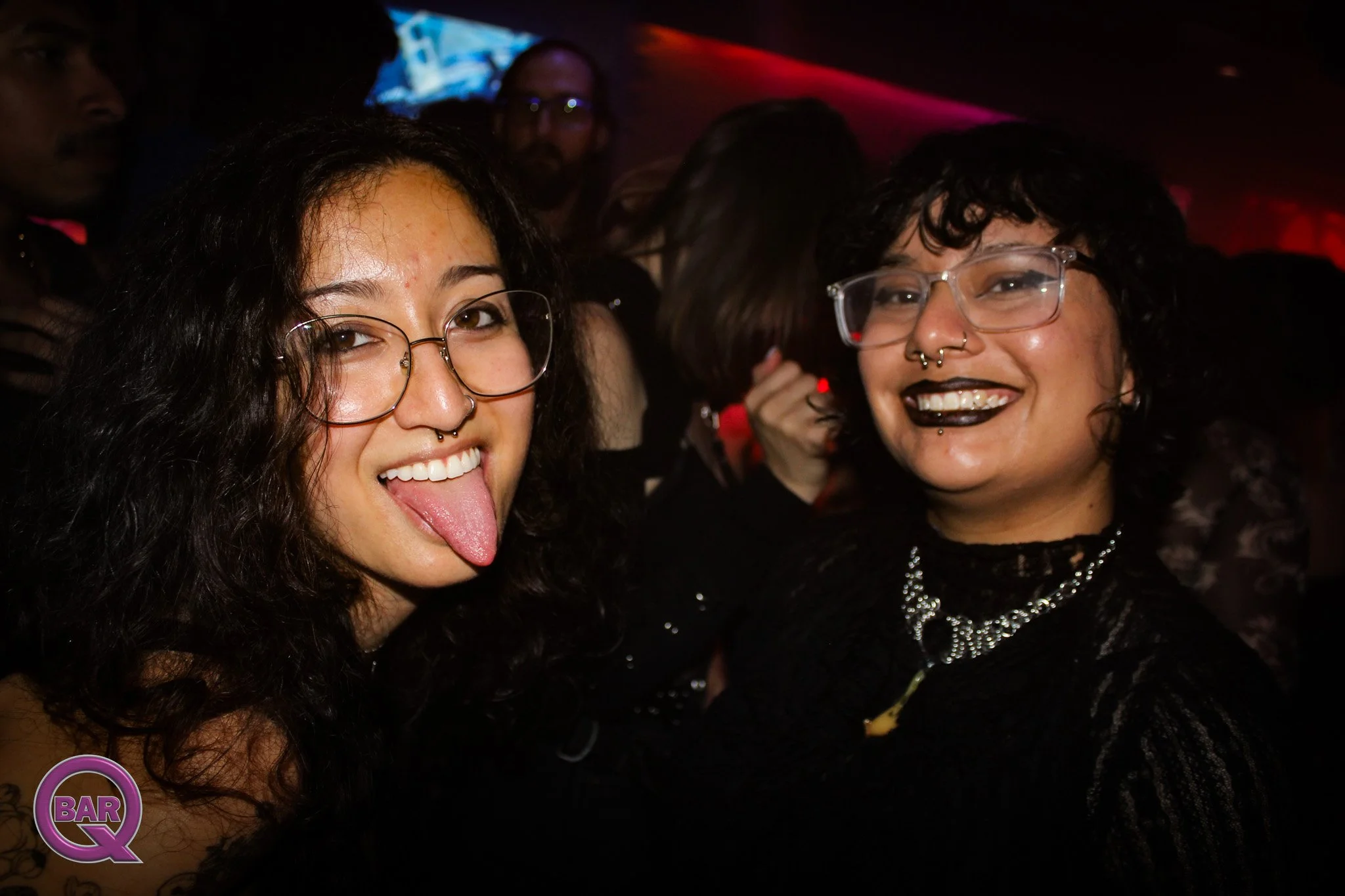 Two women smiling and posing for a photo in a dimly lit setting, possibly a nightclub or bar, with the woman on the left sticking her tongue out and the woman on the right wearing glasses and dark lipstick.