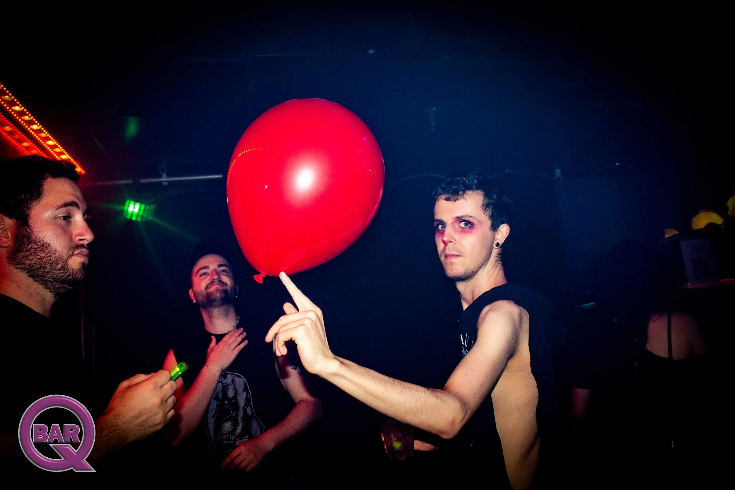 A group of young people at a party, one holding a red balloon, with colorful lighting and a neon 'BAR' sign in the background.