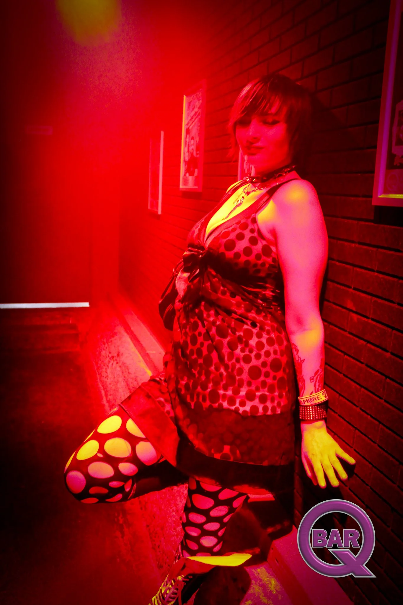 A person with short hair and light skin wearing a red and black polka dot dress, matching leggings, and red shoes, standing in a bar or nightclub with red lighting. They are holding a bag with a polka dot pattern, wearing a choker, bracelets, and a t