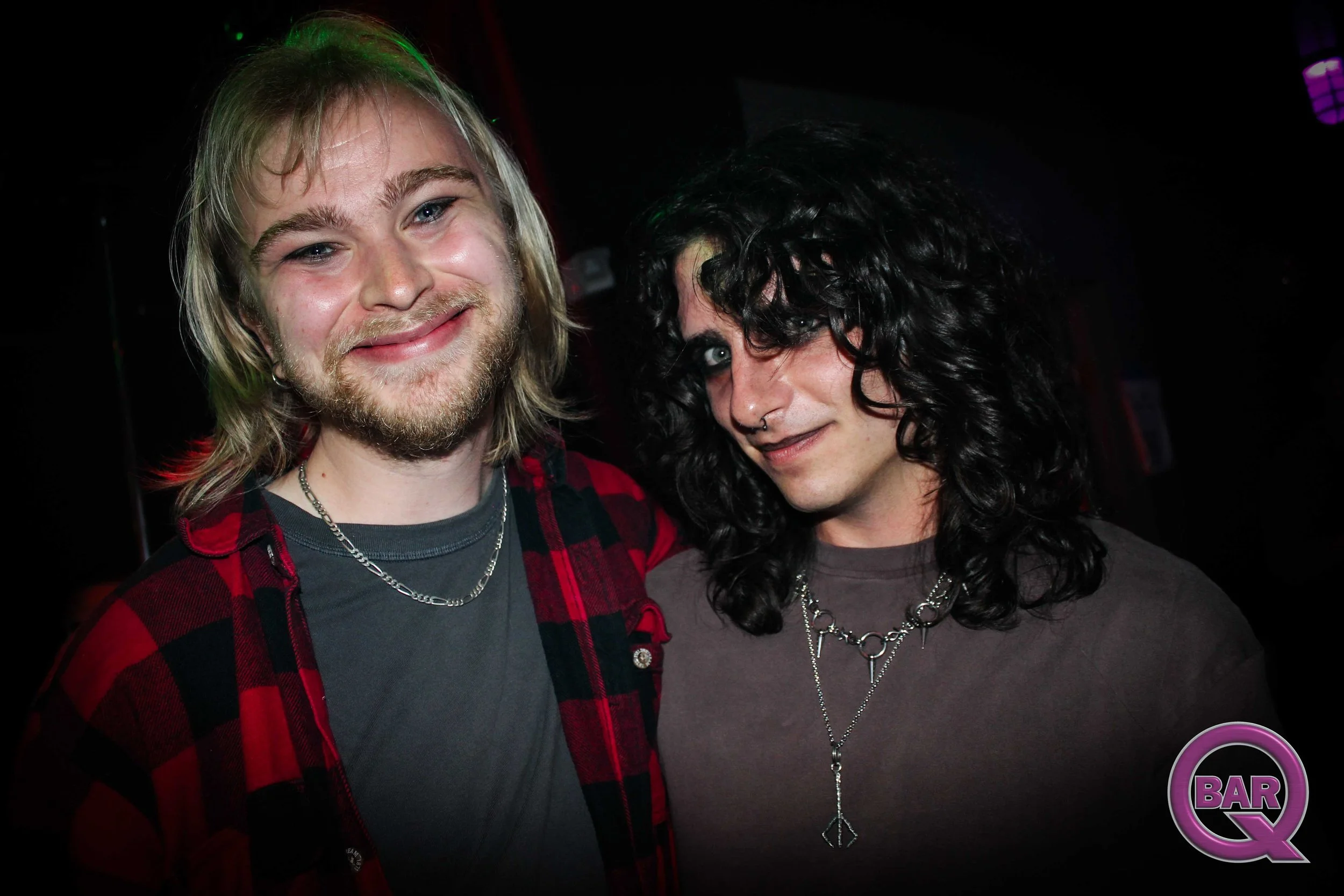 Two people smiling at a bar, one with blonde hair and a beard wearing a red flannel shirt and the other with dark curly hair and piercings wearing a gray shirt.