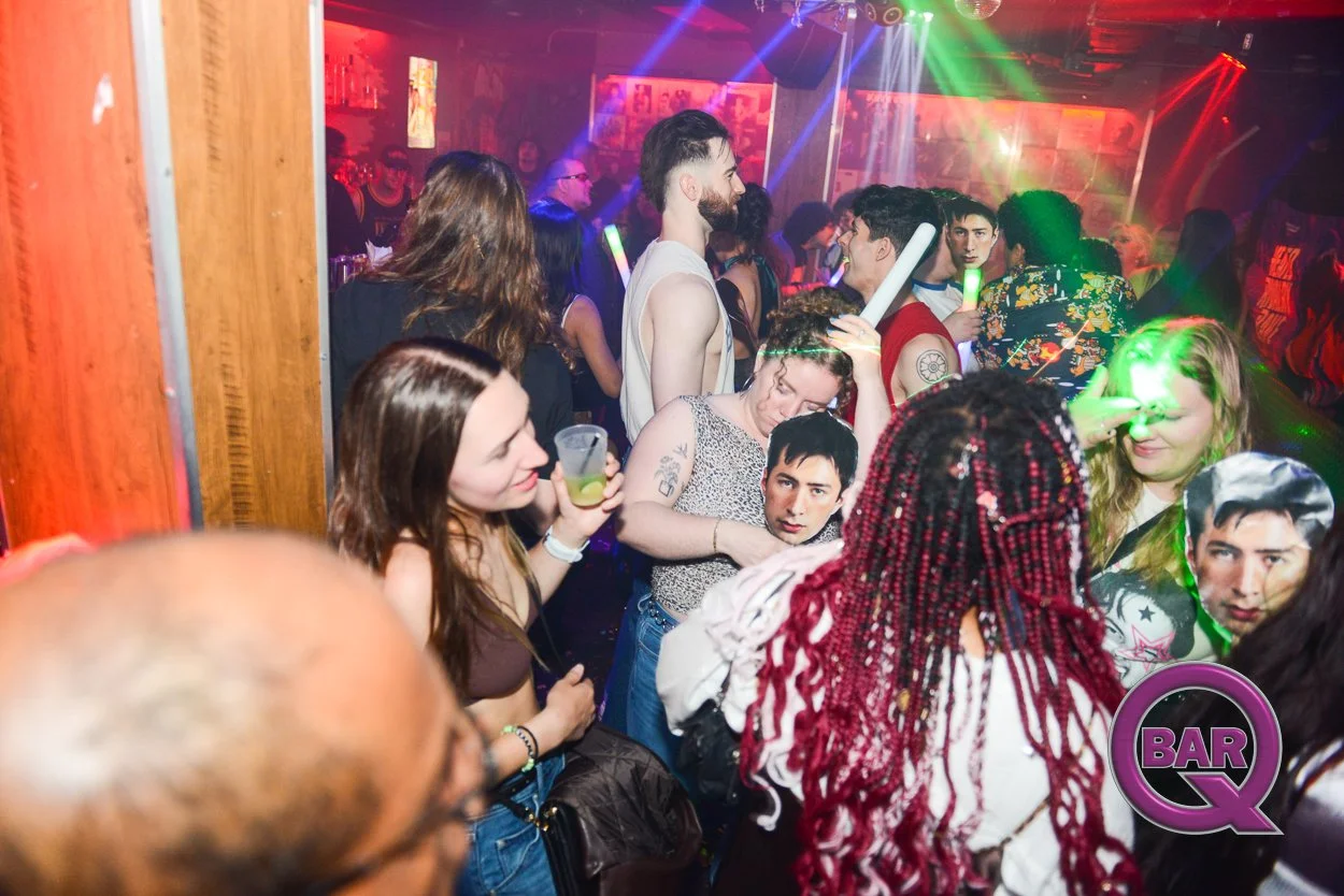 Crowd of young people dancing and socializing at a nightclub with colorful lights and a bar area in the background.
