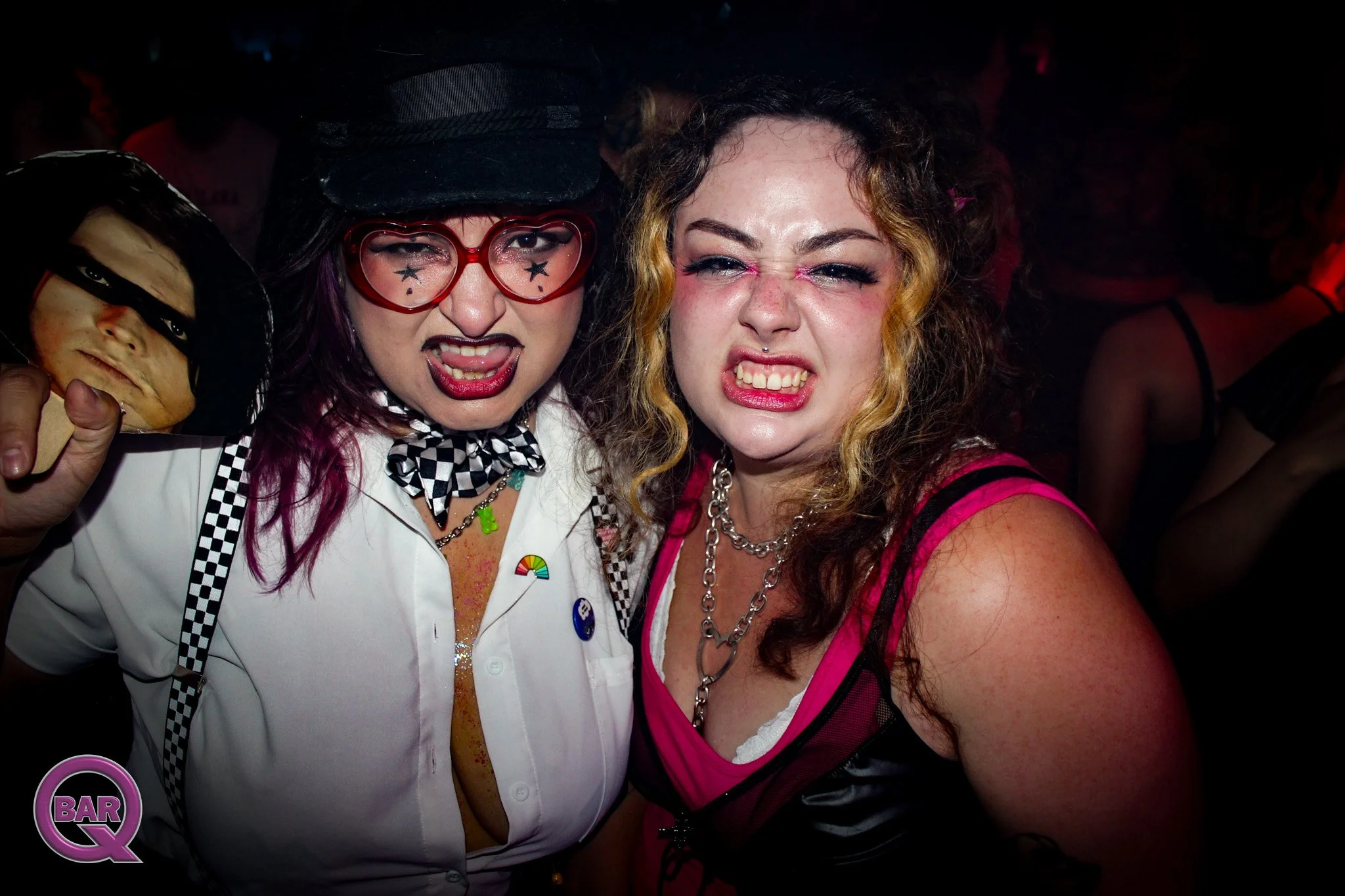 Two women at a party taking a selfie, with exaggerated facial expressions. The woman on the left has purple hair, heart-shaped glasses, face makeup with stars, and is wearing a checkered scarf and a white shirt. The woman on the right has curly hair 