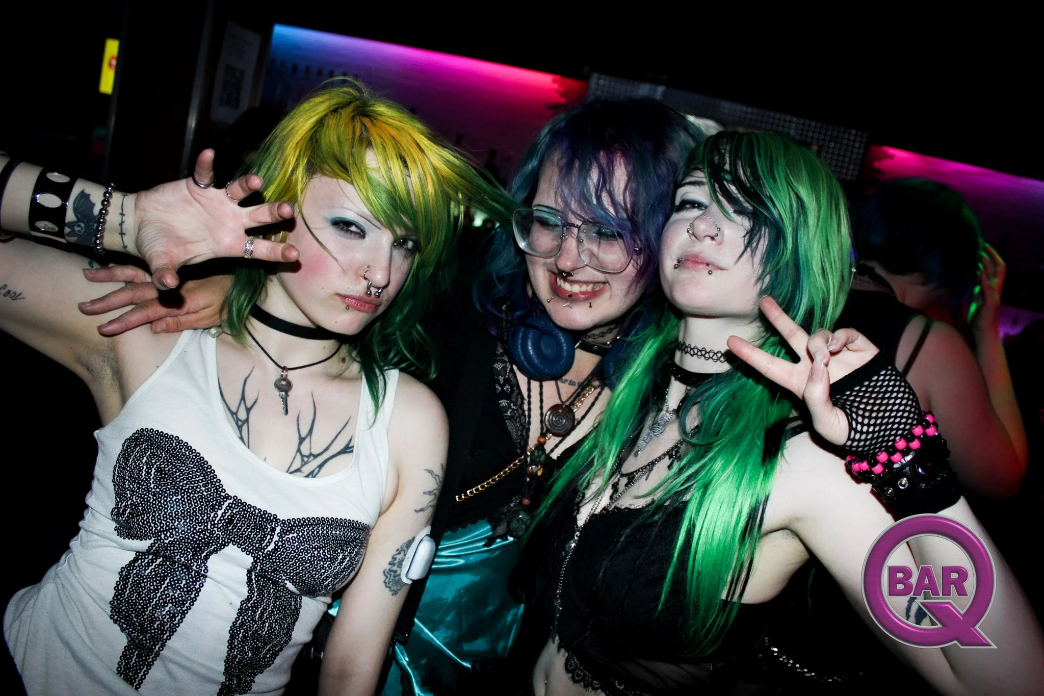 Three young women with colorful hair and multiple piercings smiling and posing together in a dark, neon-lit club.