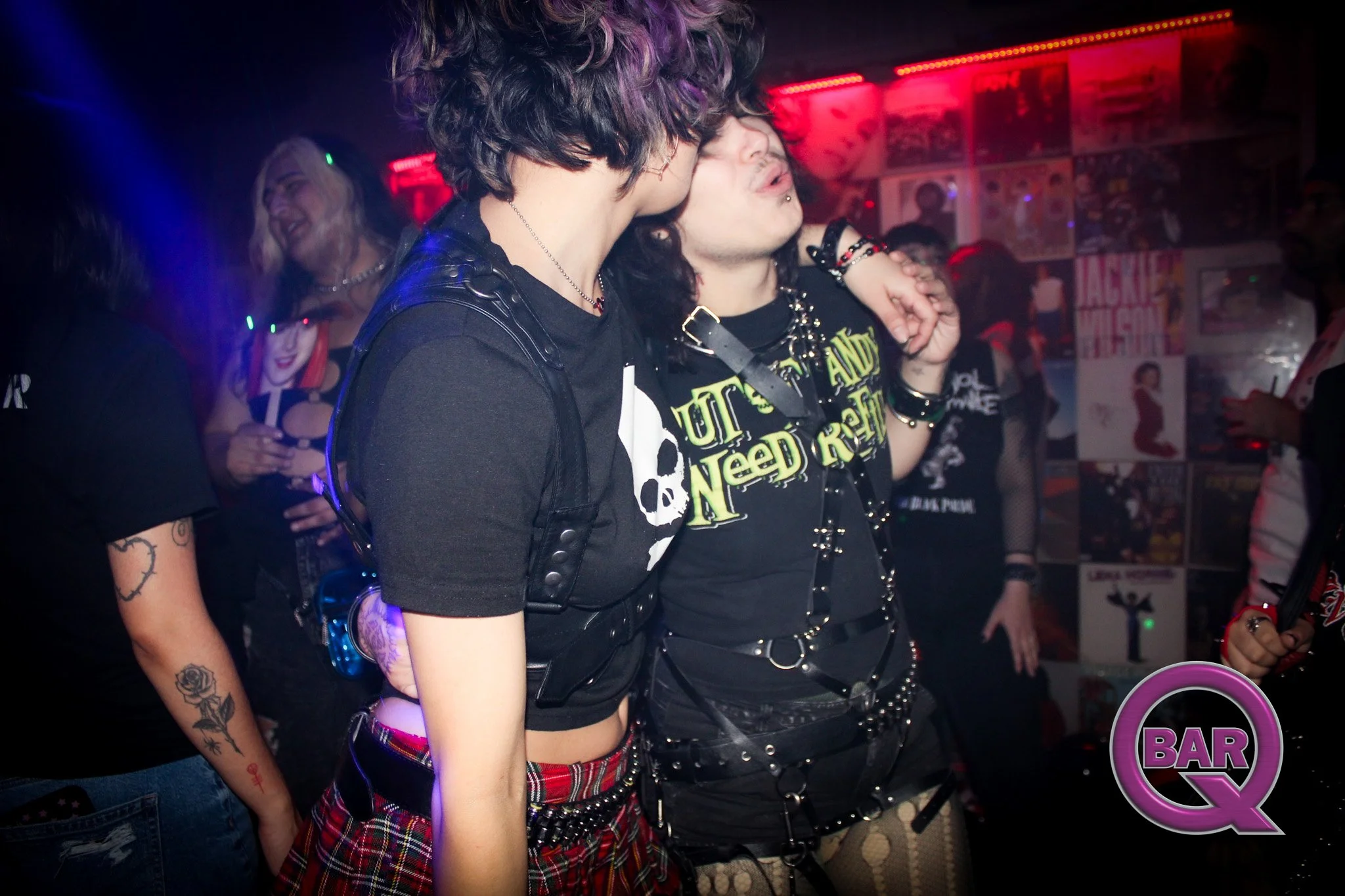 Two young people with dark, wavy hair sharing a kiss at a bar or club, surrounded by other people and posters on the wall, with colorful lighting and a vibrant atmosphere.