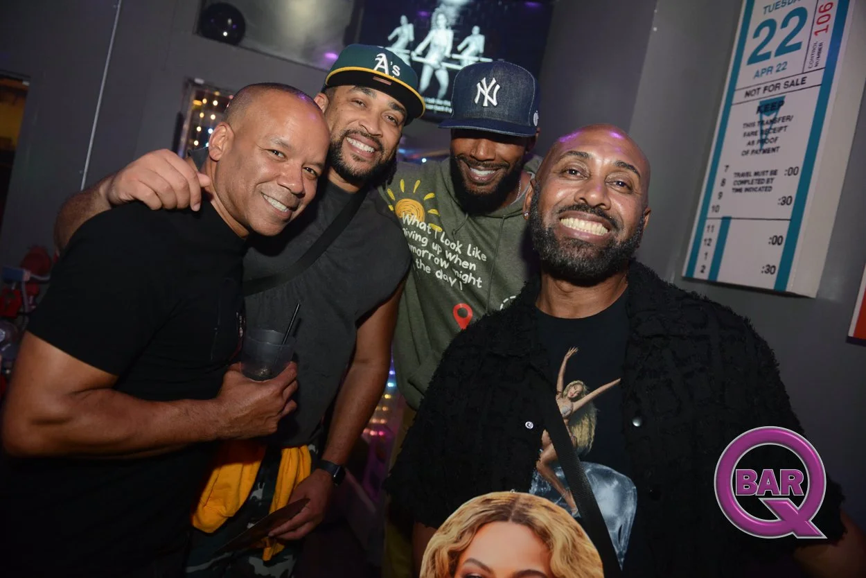 Four men smiling and posing for a photo in a bar or nightclub, with one holding a drink. There is a TV screen and a calendar on the wall behind them, and a pink and purple 'Bar Q' logo in the bottom right corner.