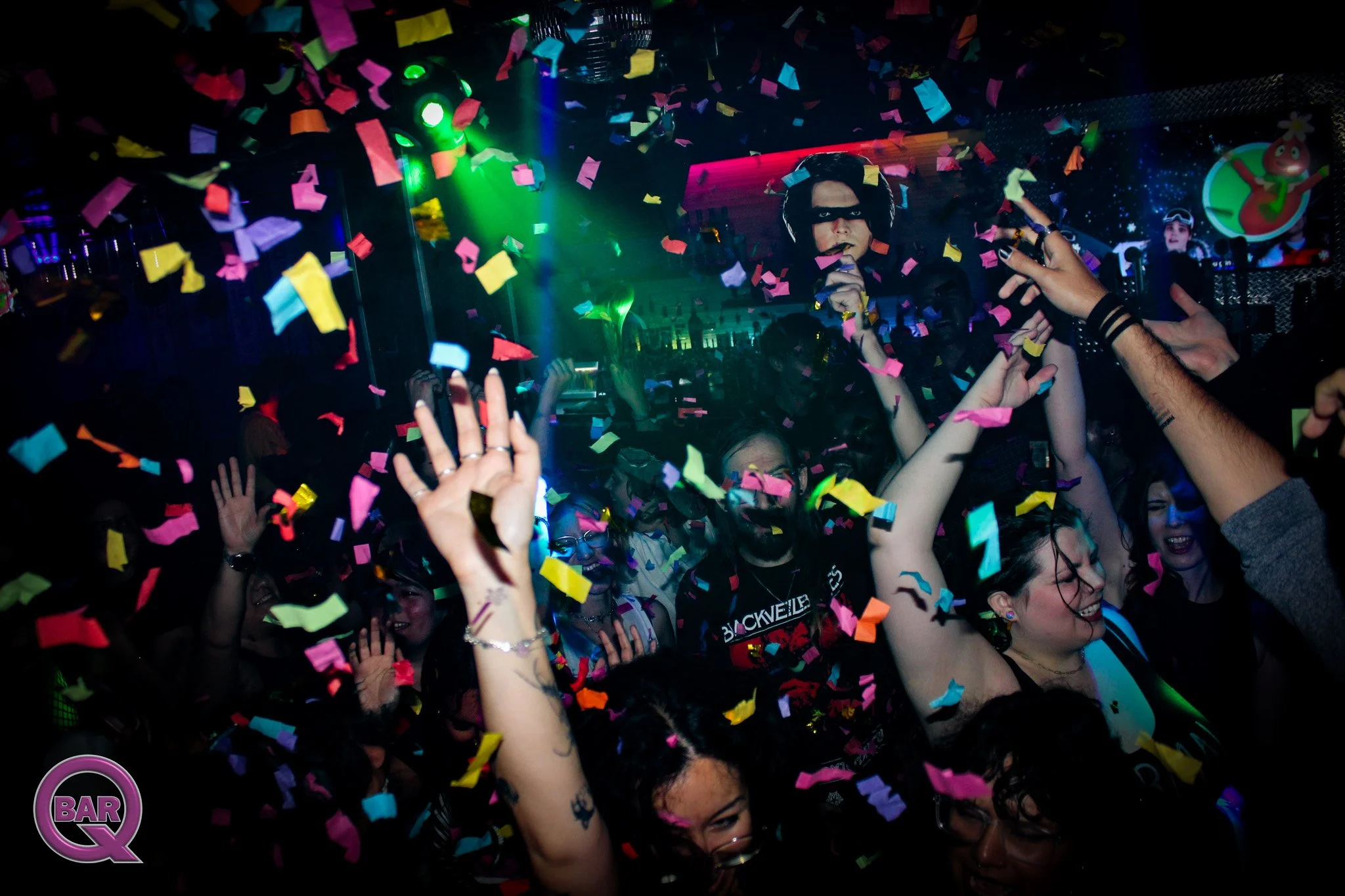 Crowd of people at a party or concert with colorful confetti falling, some raising their hands, and a woman in the center holding a microphone, with one wearing glasses and tattoos on her arm, in a lively, dark club setting with neon lights and scree