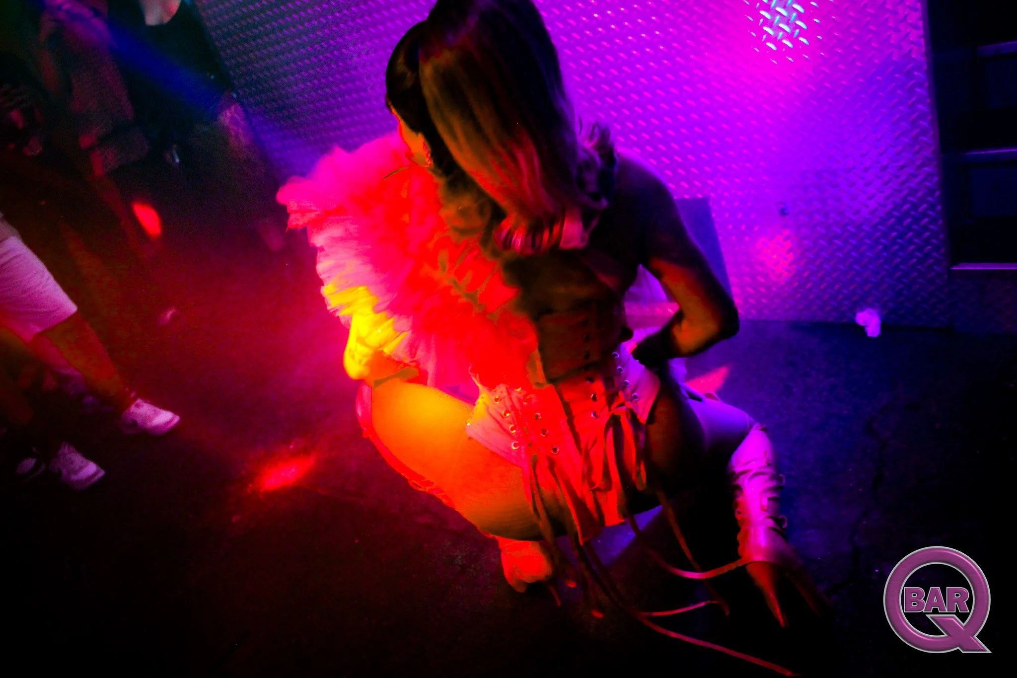 Person sitting at a nightclub or bar, wearing a black top and white shorts, with colorful neon lights reflecting on the walls and floor, and people standing nearby.