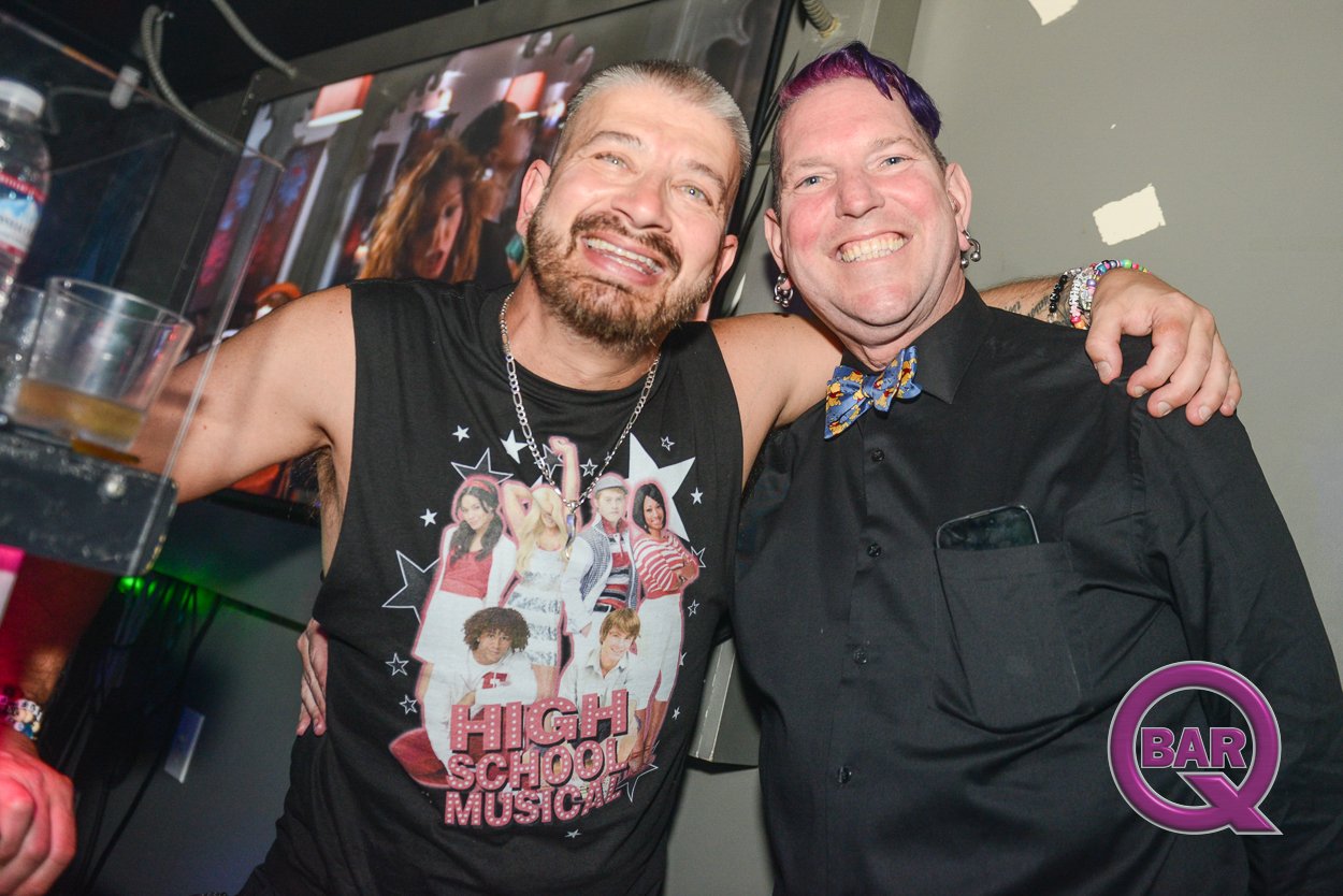 Two smiling men with arm around each other in a bar or club setting, one wearing a sleeveless High School Musical t-shirt and the other in a black shirt with a colorful bow tie, both showing excitement and happiness.