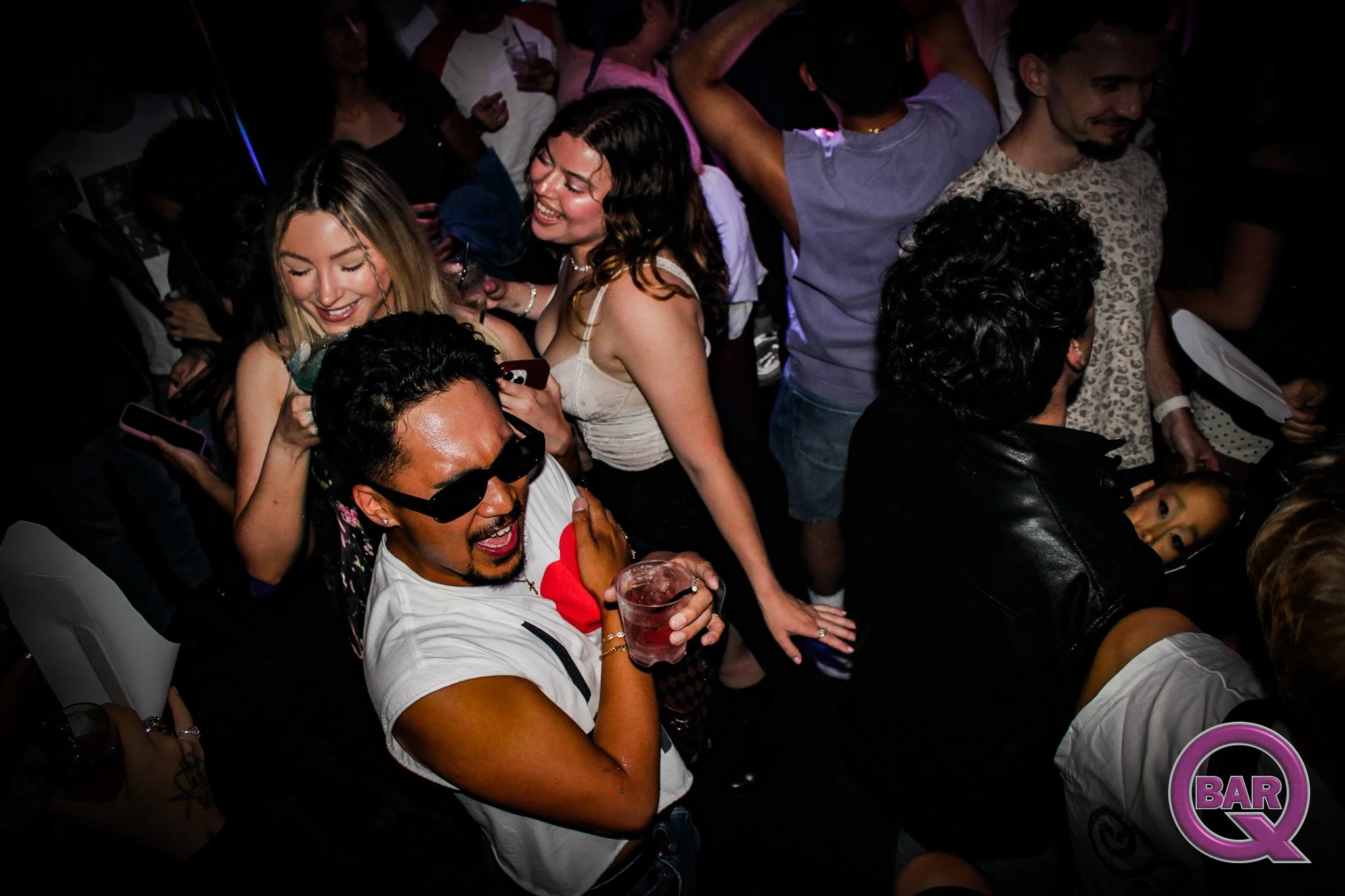 A group of people at a lively party or nightclub, smiling and enjoying drinks, with some using their phones, and a neon sign in the background.