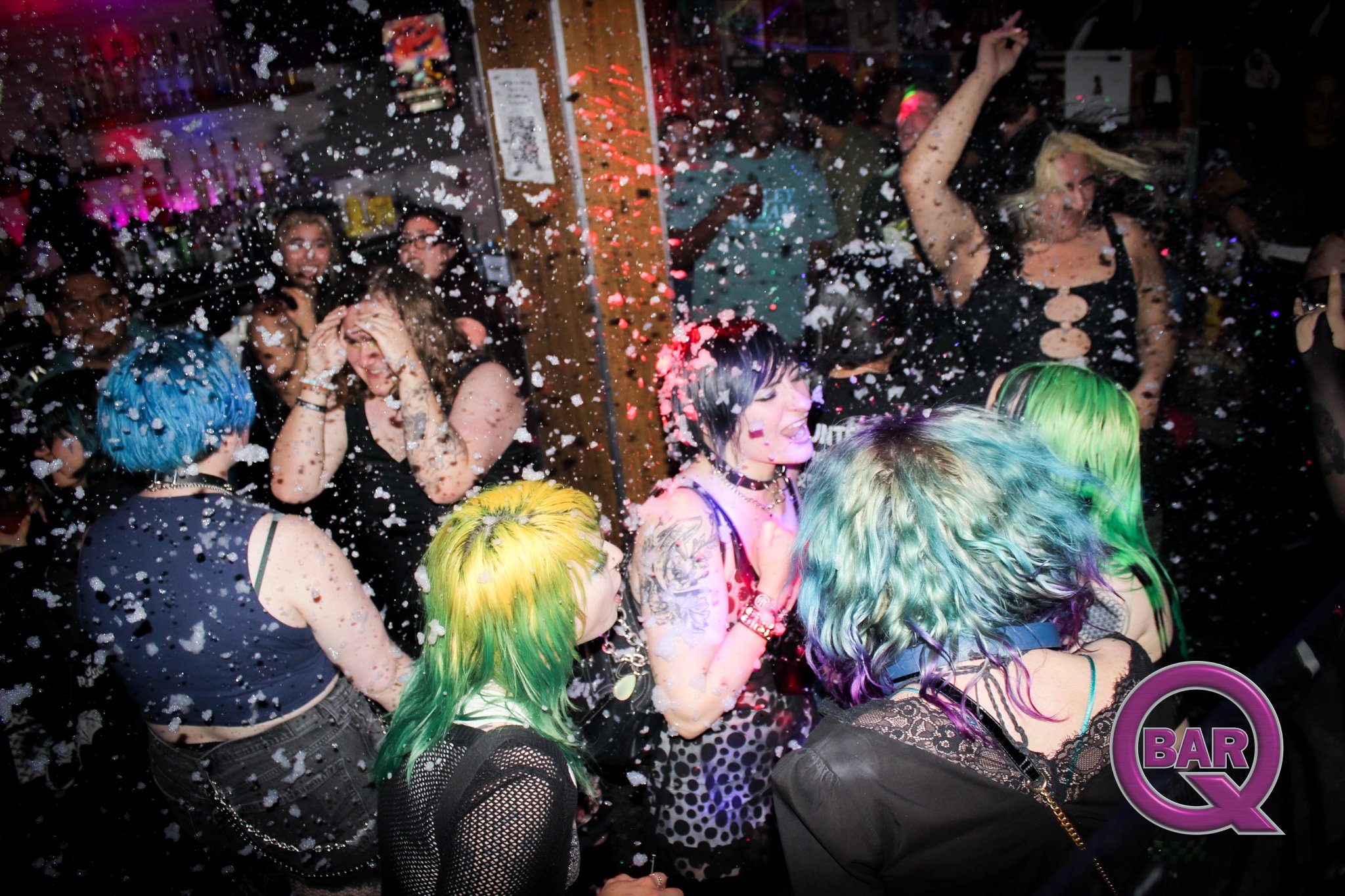 People dancing and having fun at a nightclub party with confetti falling around them. Bright colorful hair, tattoos, and punk or goth fashion styles are visible.