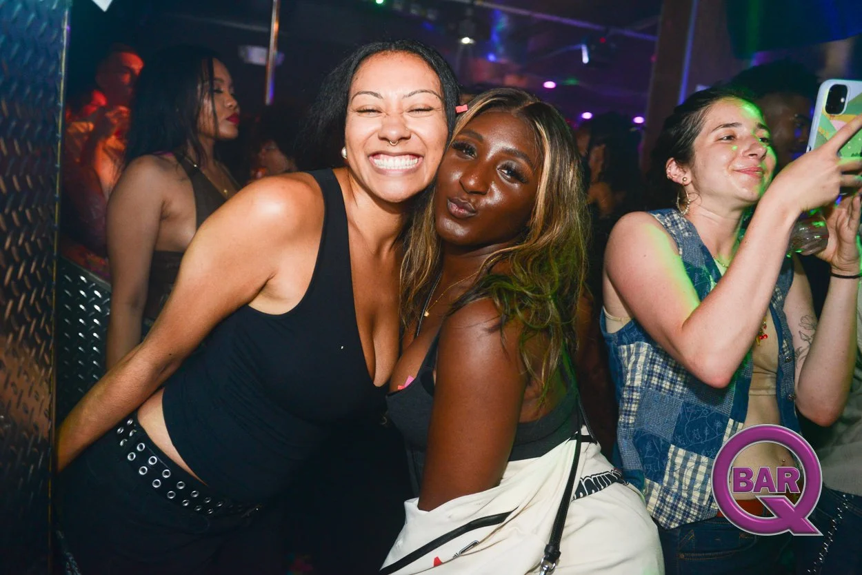 Two women smiling and posing for a photo at a lively bar or club with colorful lighting and other people in the background. One woman has curly hair and a septum piercing, and the other has wavy hair and is making a pouty face. A third woman on the r