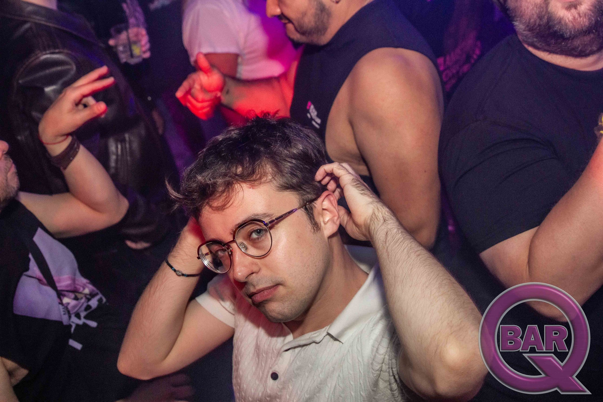 A group of people dancing and socializing in a bar or nightclub with purple lighting. Prominent in the image is a young man wearing glasses, a white shirt, and a focused expression, holding his head with one hand. The background features other people