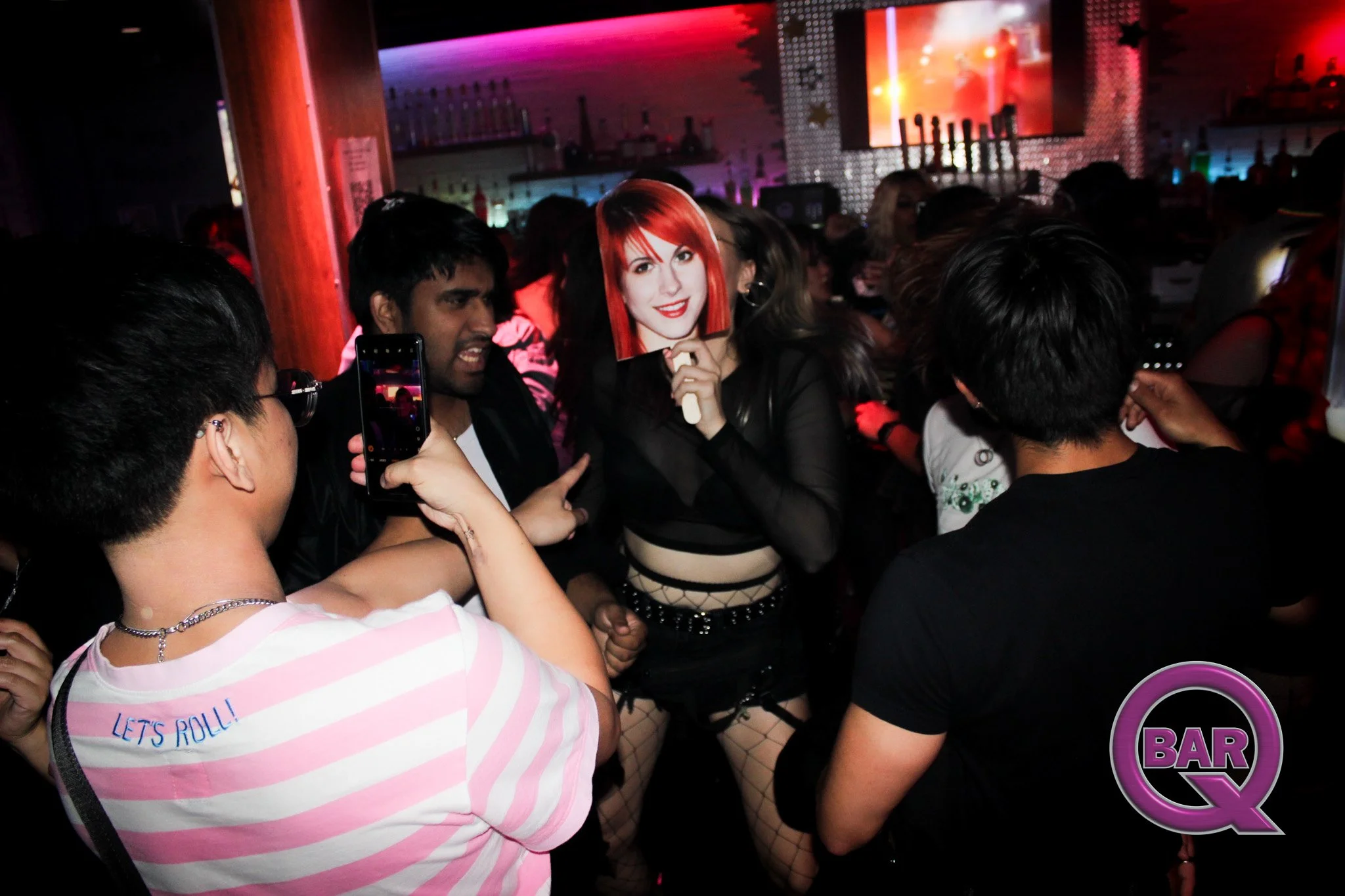 A crowded party scene with people dancing and taking photos, a woman holding a mask of a woman with red hair and red lipstick, and a bar sign in the background with colorful lighting.