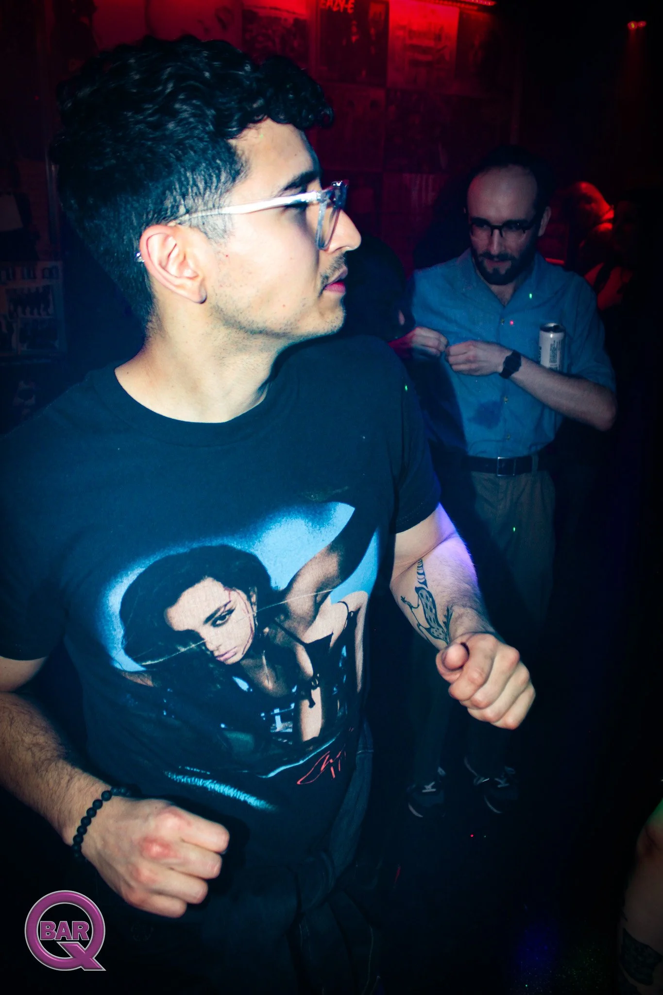 A man with dark curly hair and glasses standing in a dimly lit bar or club with red and blue lighting. He is wearing a graphic T-shirt of a woman and is holding something in his left hand. Behind him, another man with glasses and a beard is holding a