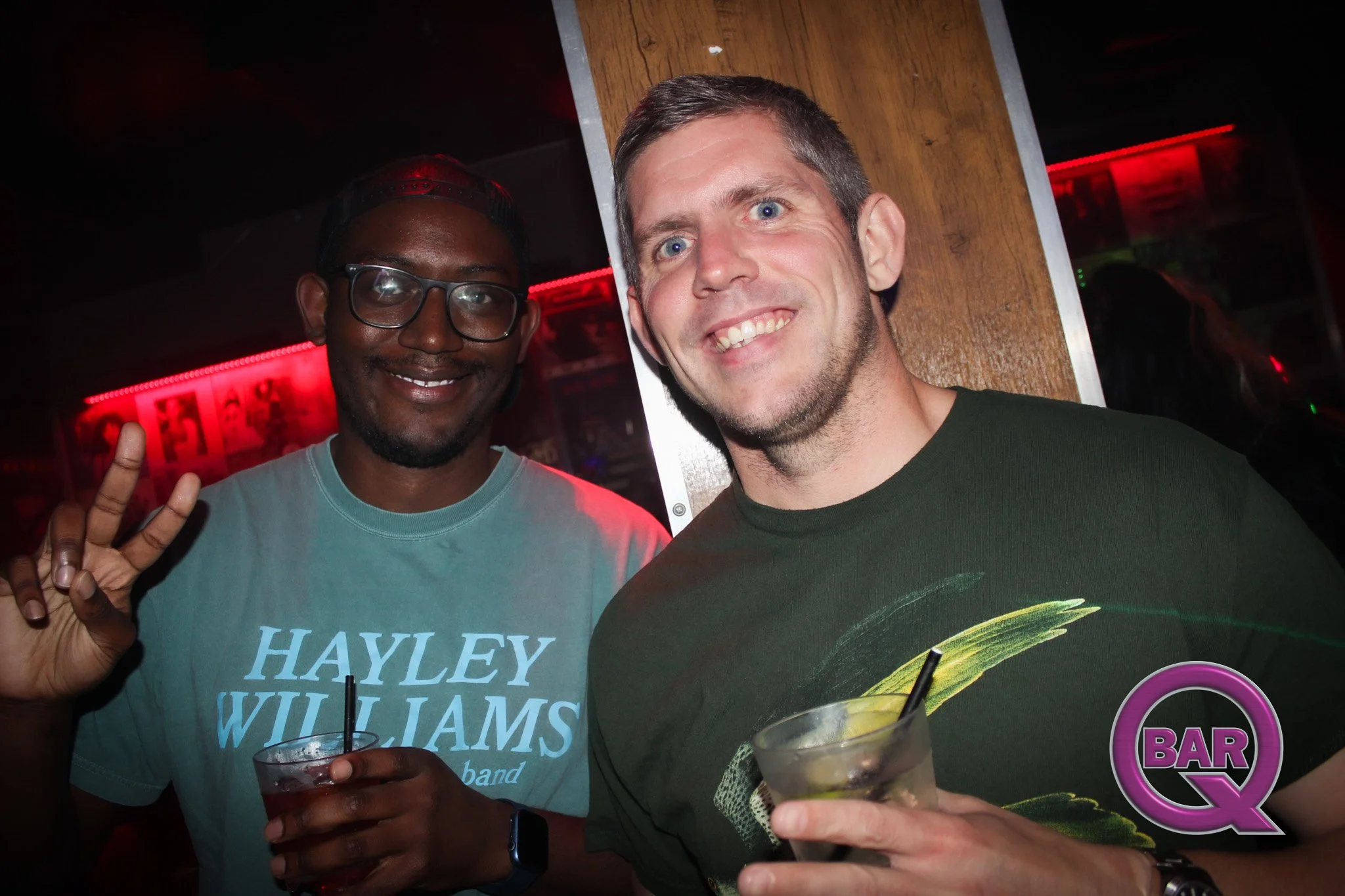 Two men smiling and holding drinks at a bar, one wearing glasses and a teal T-shirt, the other with short hair and a dark T-shirt, with a wooden column behind them and red lighting in the background.