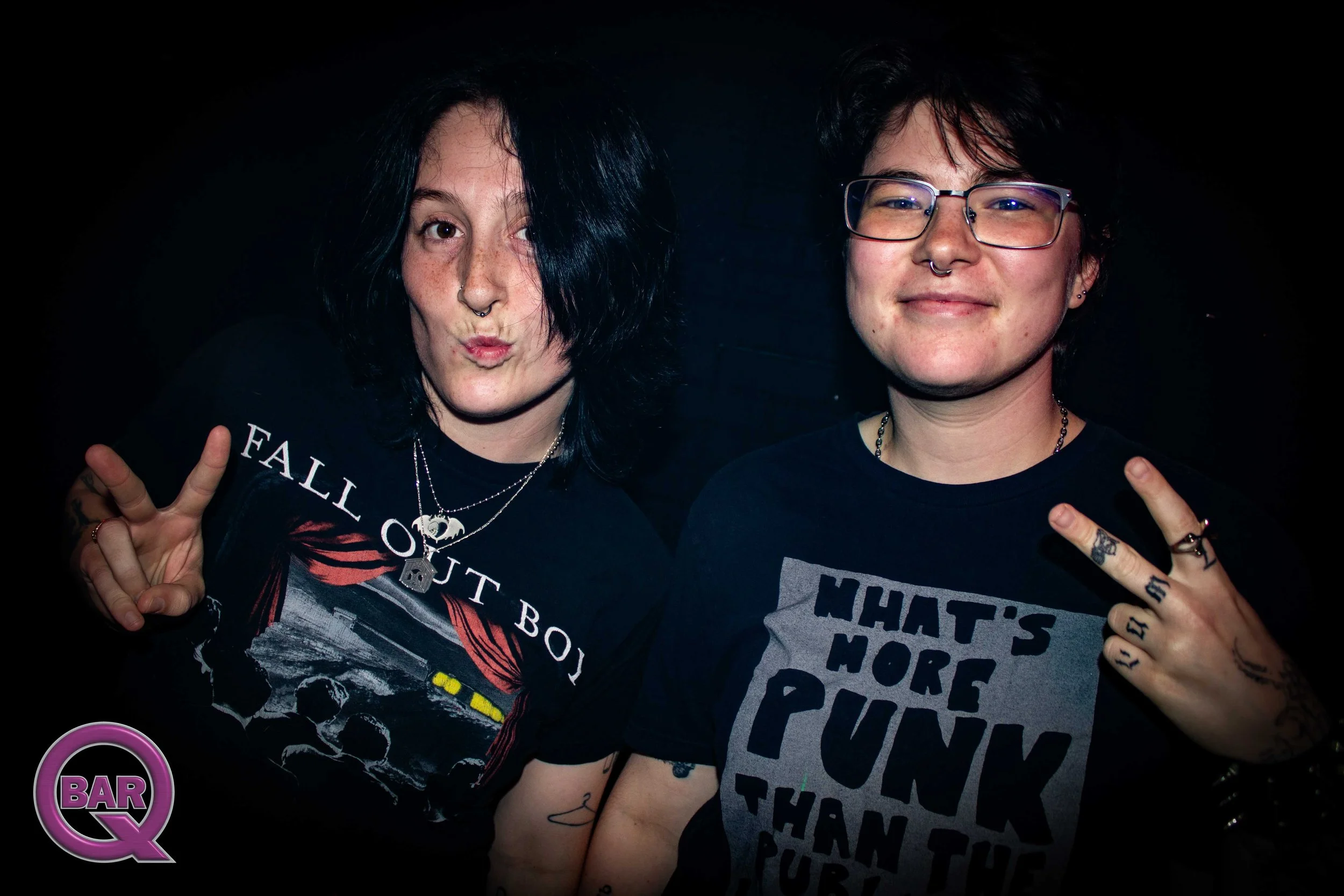 Two people with dark hair and glasses, making peace signs, posing for a photo at a bar or club.