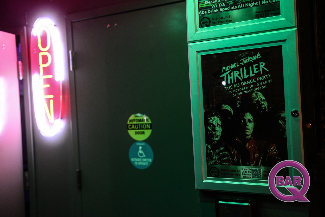 Neon 'OPEN' sign, posters for Michael Jackson's Thriller dance party, and various stickers on a dark wall outside a bar in San Francisco.