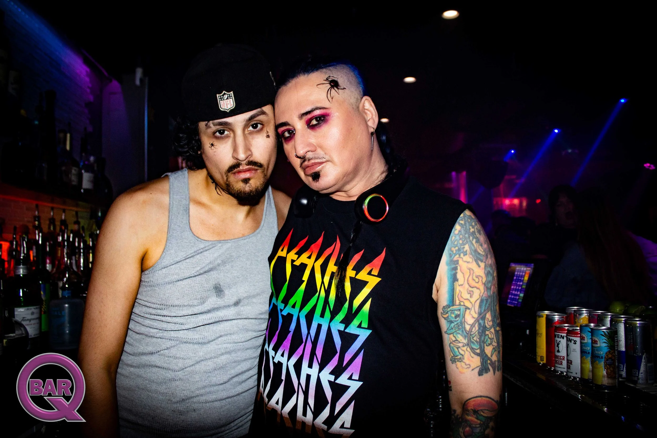 Two men stand close together in a dimly lit bar or club. The man on the left has curly hair, facial hair, and wears a gray tank top and a black cap with an NFL logo. The man on the right has colored hair, makeup, tattoos, and wears a colorful, sleeve