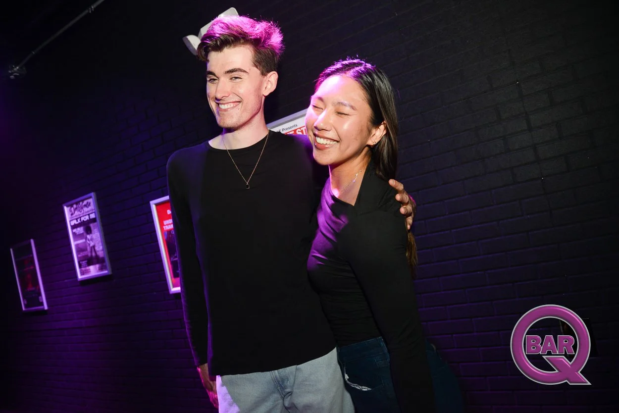 A young man and woman smiling and posing together in a dimly lit bar with purple and pink lighting, black brick wall in the background, and framed posters on the wall.