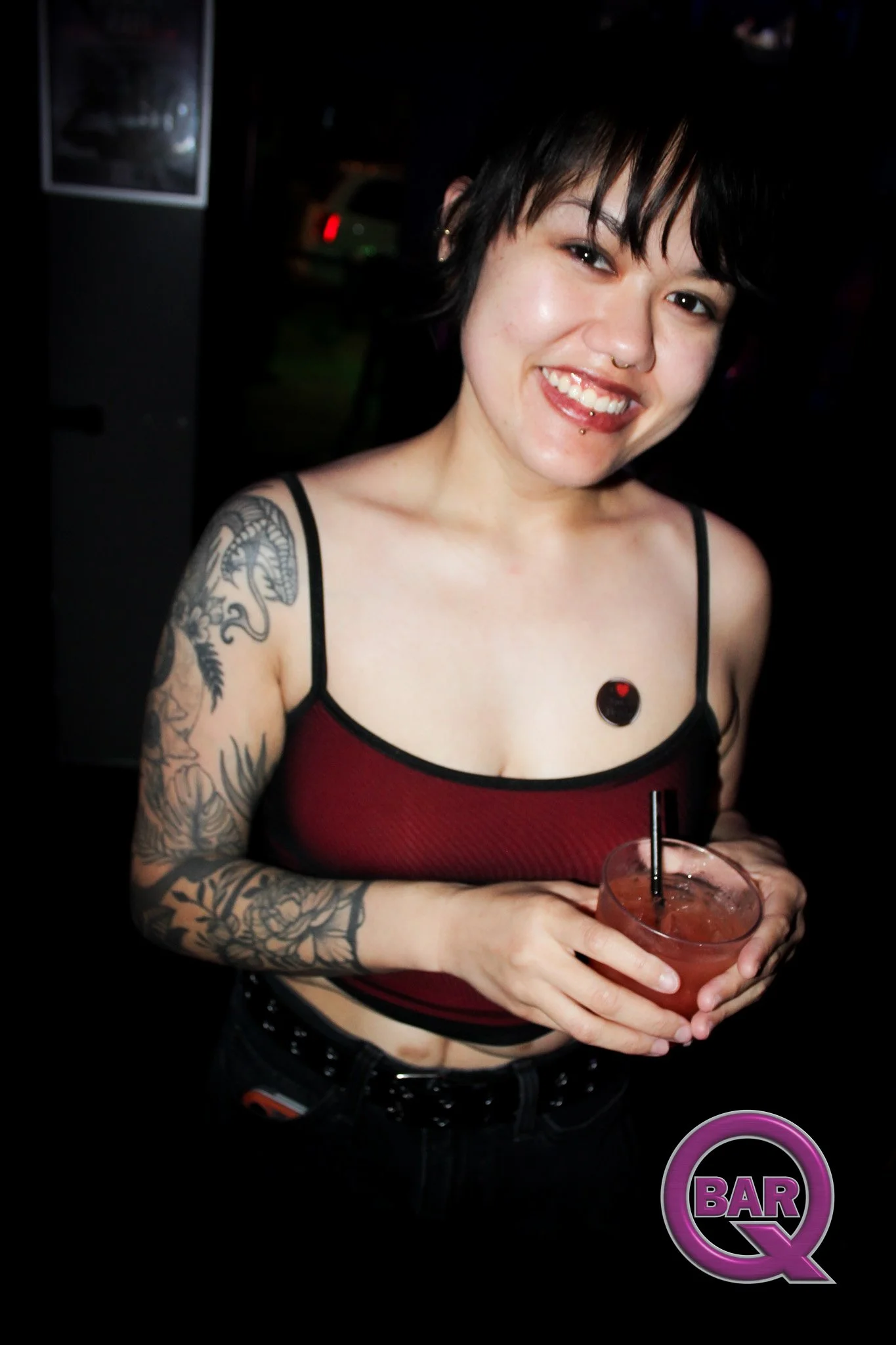 A young woman with short black hair, tattoos on her right arm, and piercings on her nose and lip, holding a glass of drink. She is smiling at the camera, wearing a red and black sleeveless top, and standing in front of a nightclub or bar sign.