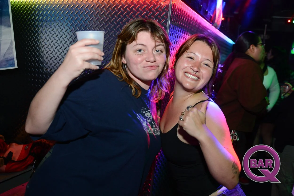 Two women smiling and enjoying themselves at a bar with colorful lighting, one holding a drink and the other giving a thumbs-up.