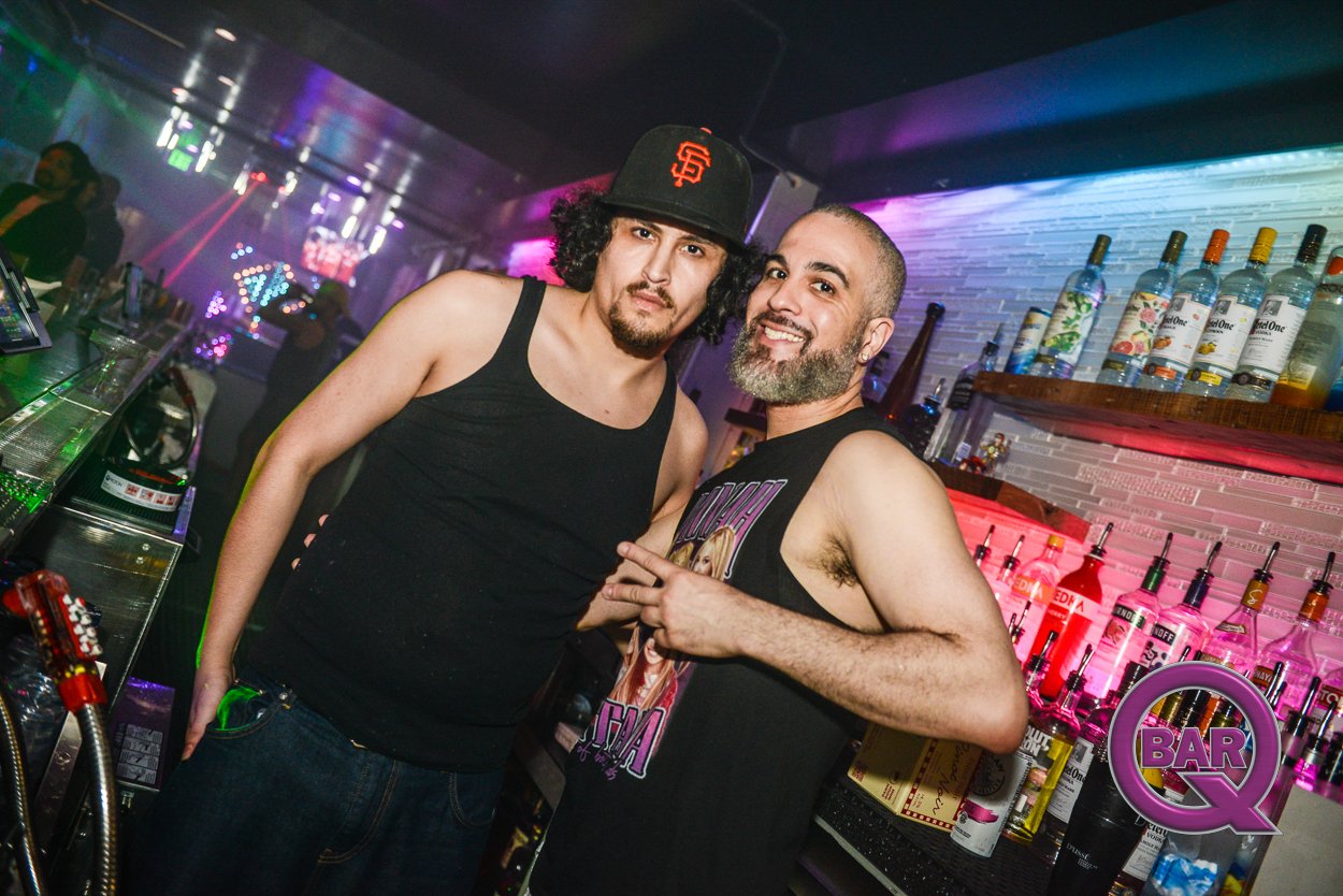 Two men posing at a bar. One is wearing a black tank top and a San Francisco Giants cap, the other is wearing a Nirvana tank top and has a bearded face. The bar background has liquor bottles and colorful lighting.