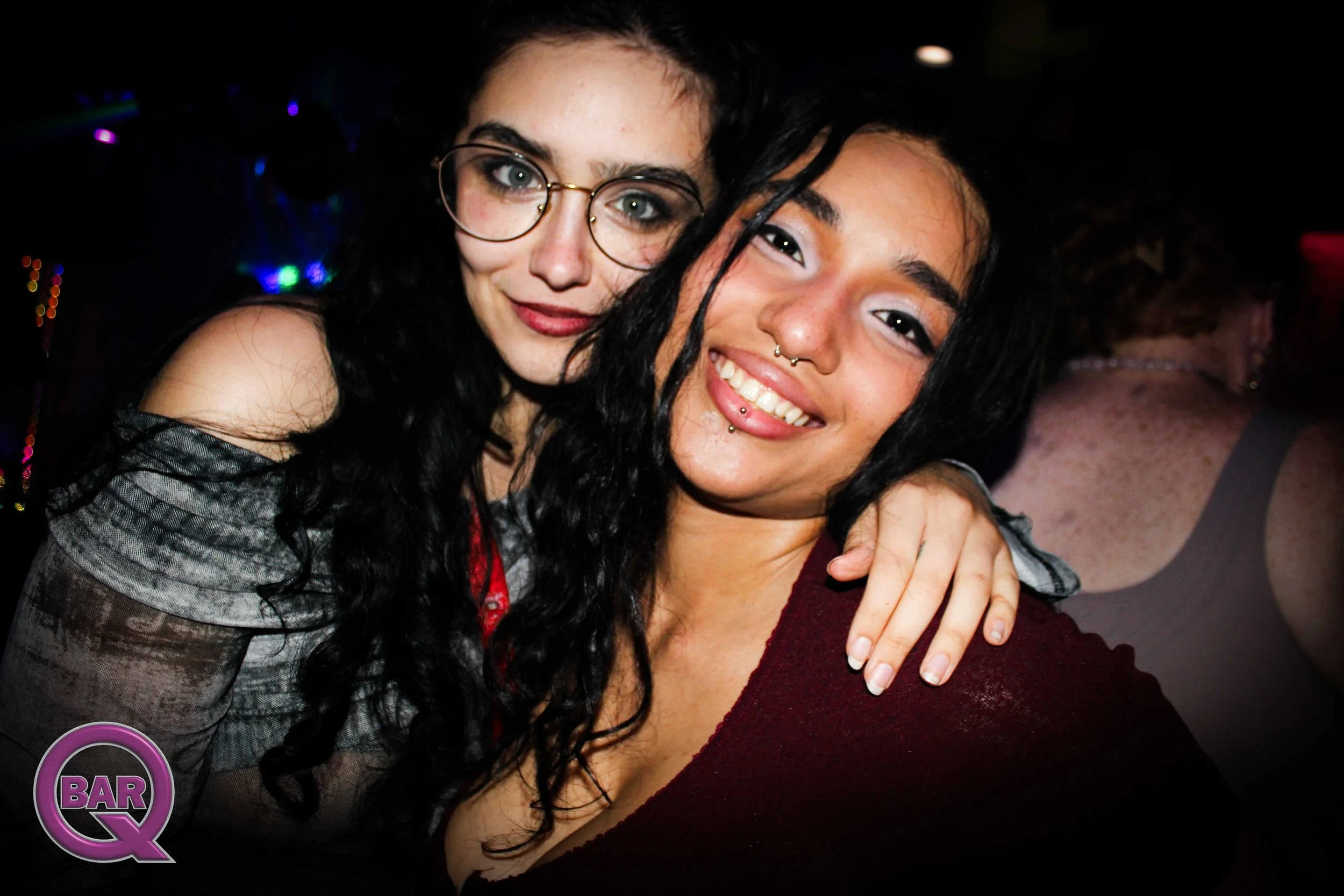 Two women with dark hair smiling and hugging at a bar or club, with a dark background and colorful lights.