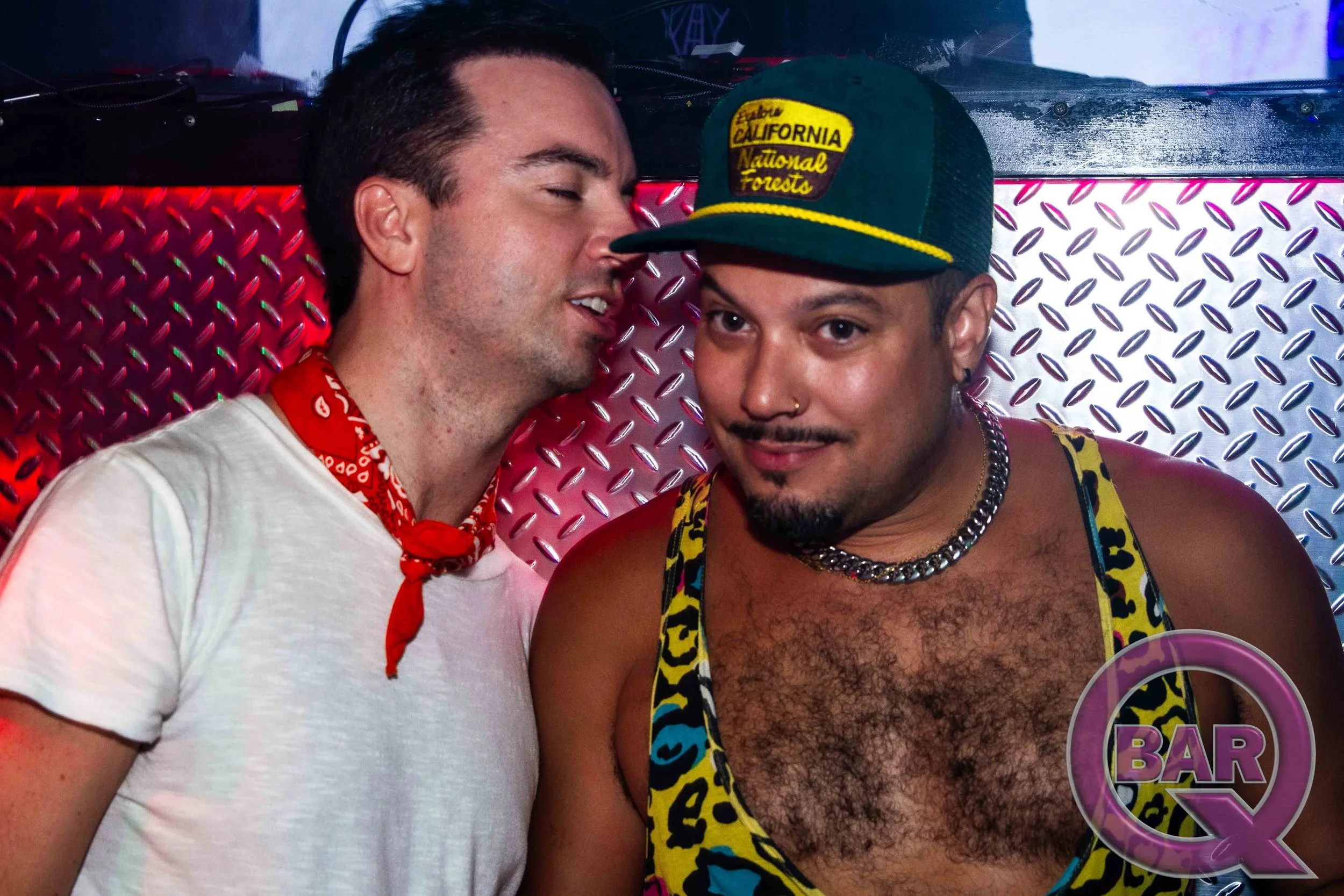 Two men close together in a nightclub or bar setting, one wearing a white t-shirt and a red bandana, the other in a colorful tank top and a dark green cap with yellow lettering. They are in front of a red metallic background with diamond patterns, wi