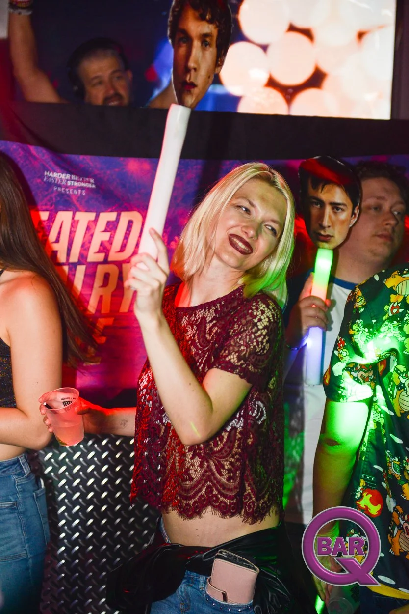 A woman with blonde hair in a lace red top dancing with a drink in her hand at a nightclub, surrounded by other people, neon lights, and a large poster of a person behind her.