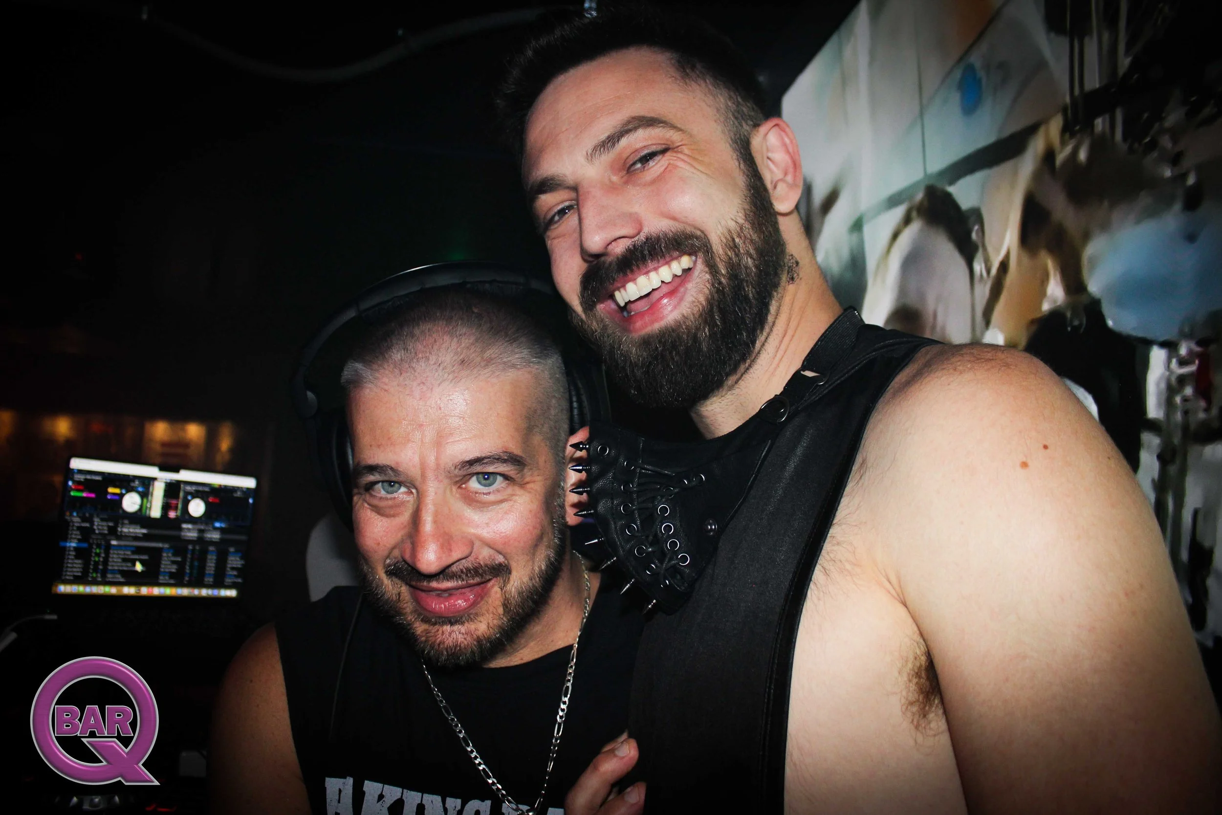 Two men smiling at a party, one with a beard and the other wearing headphones, with a DJ setup in the background.