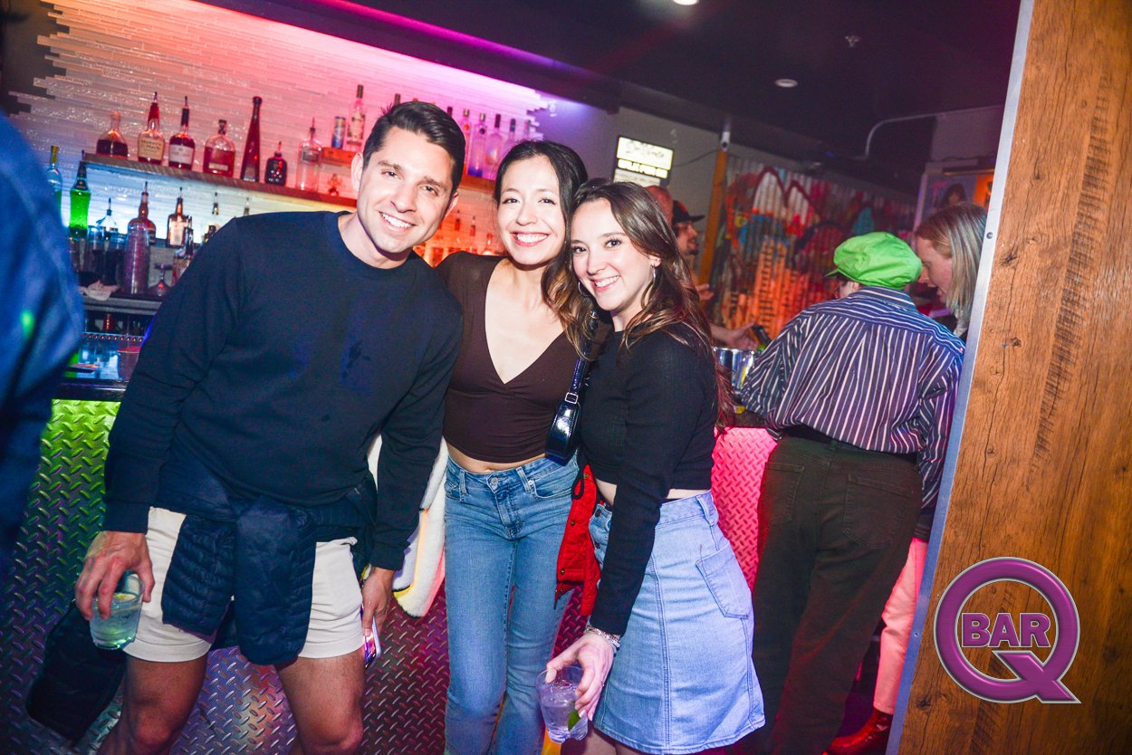 Three smiling young adults, two women and one man, pose together in a bar with a colorful illuminated background, holding drinks in their hands.