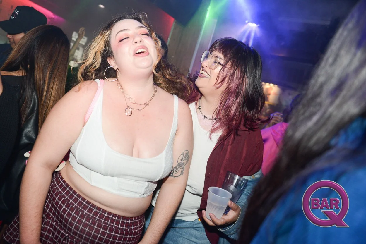 Two women laughing and enjoying themselves at a nightclub or bar, with colorful lights and other patrons in the background.