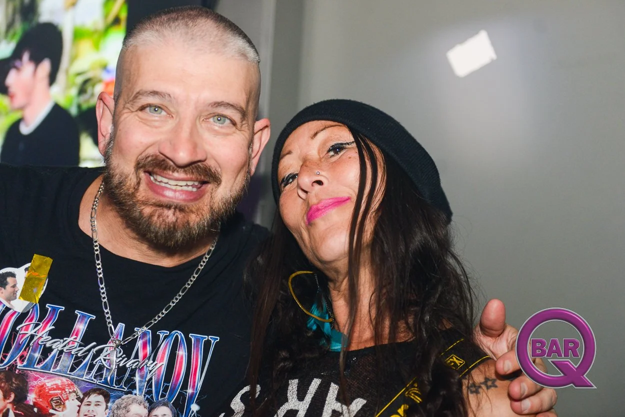 A man and woman smiling and posing closely together, with the man having blonde hair and a beard, and the woman wearing a black hat and bright pink lipstick.