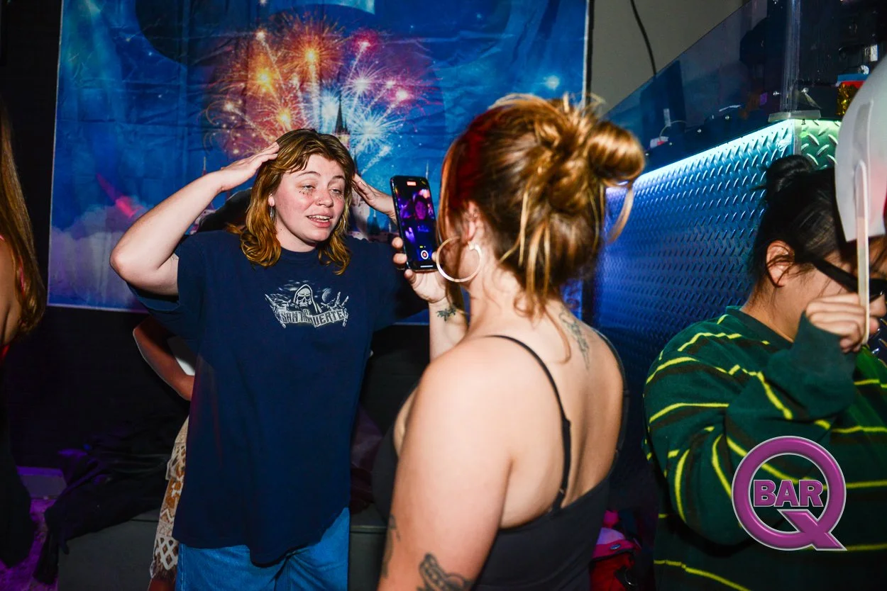 Two women at a bar, one with red hair taking a photo or video of the other, with a colorful fireworks display in the background. The woman being photographed has light skin, brown hair, and is wearing a dark T-shirt, while the woman taking the photo 