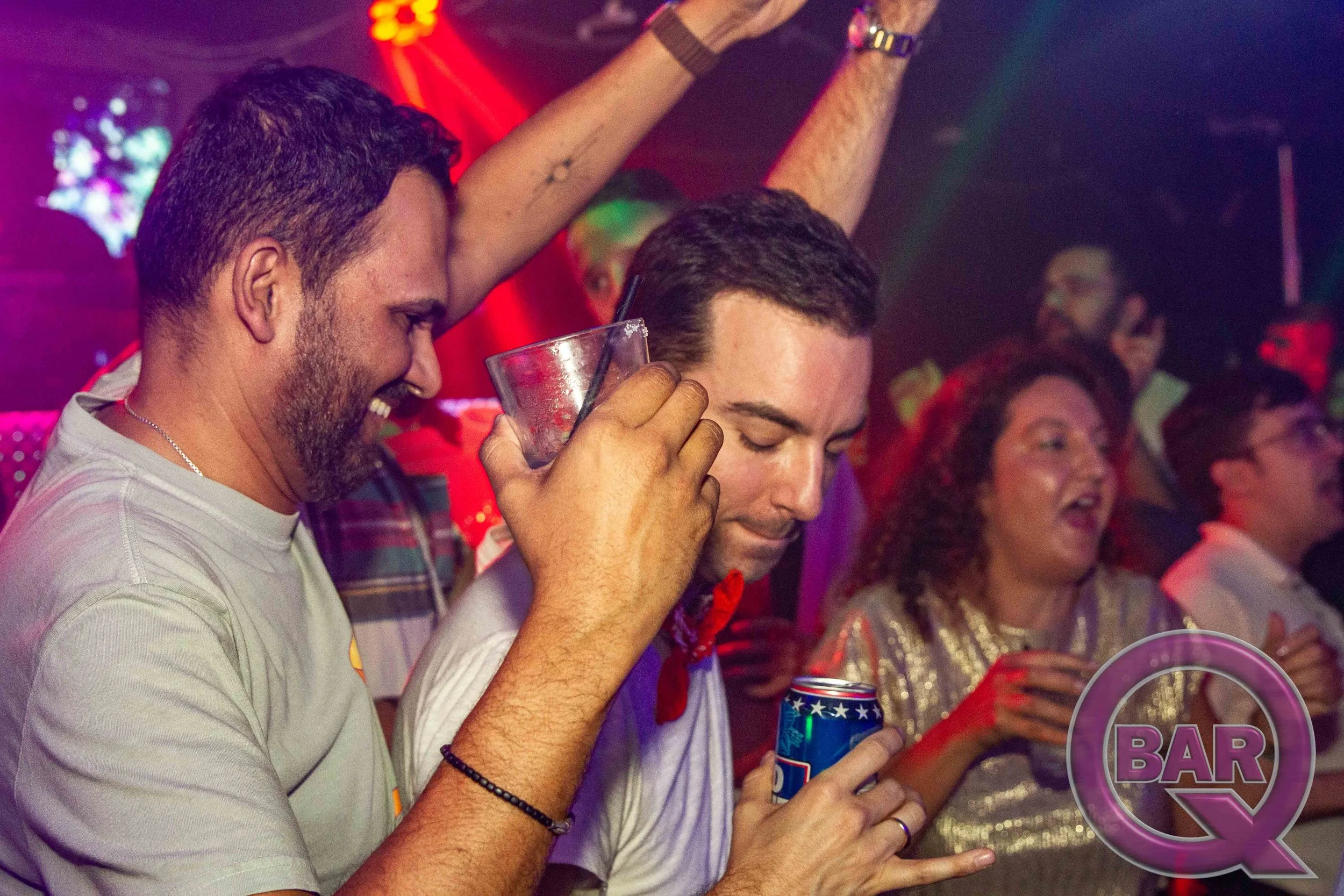 Group of diverse people at an indoor party or nightclub, smiling and holding drinks, with colorful lights and a neon sign in the background.