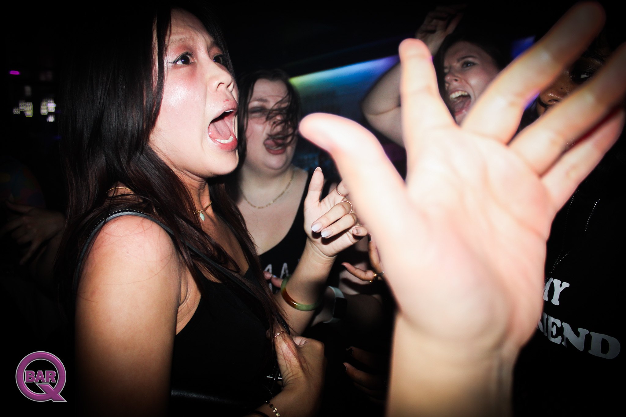 Group of women at a nightclub, singing and dancing, with one woman in the foreground with long dark hair, wearing jewelry, and others in the background with happy expressions.