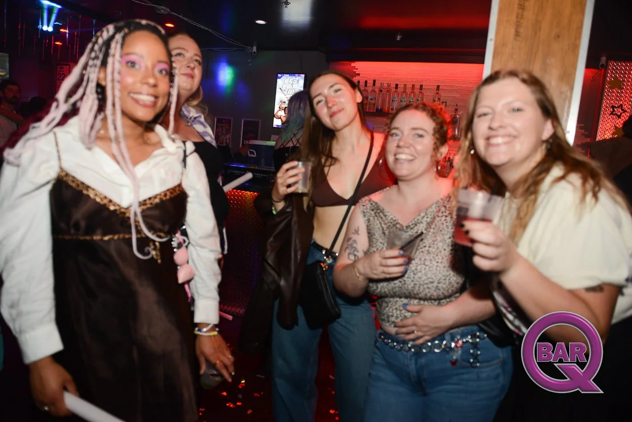 Group of five diverse women smiling and enjoying drinks at a bar or nightclub with colorful lights and a bar counter in the background.